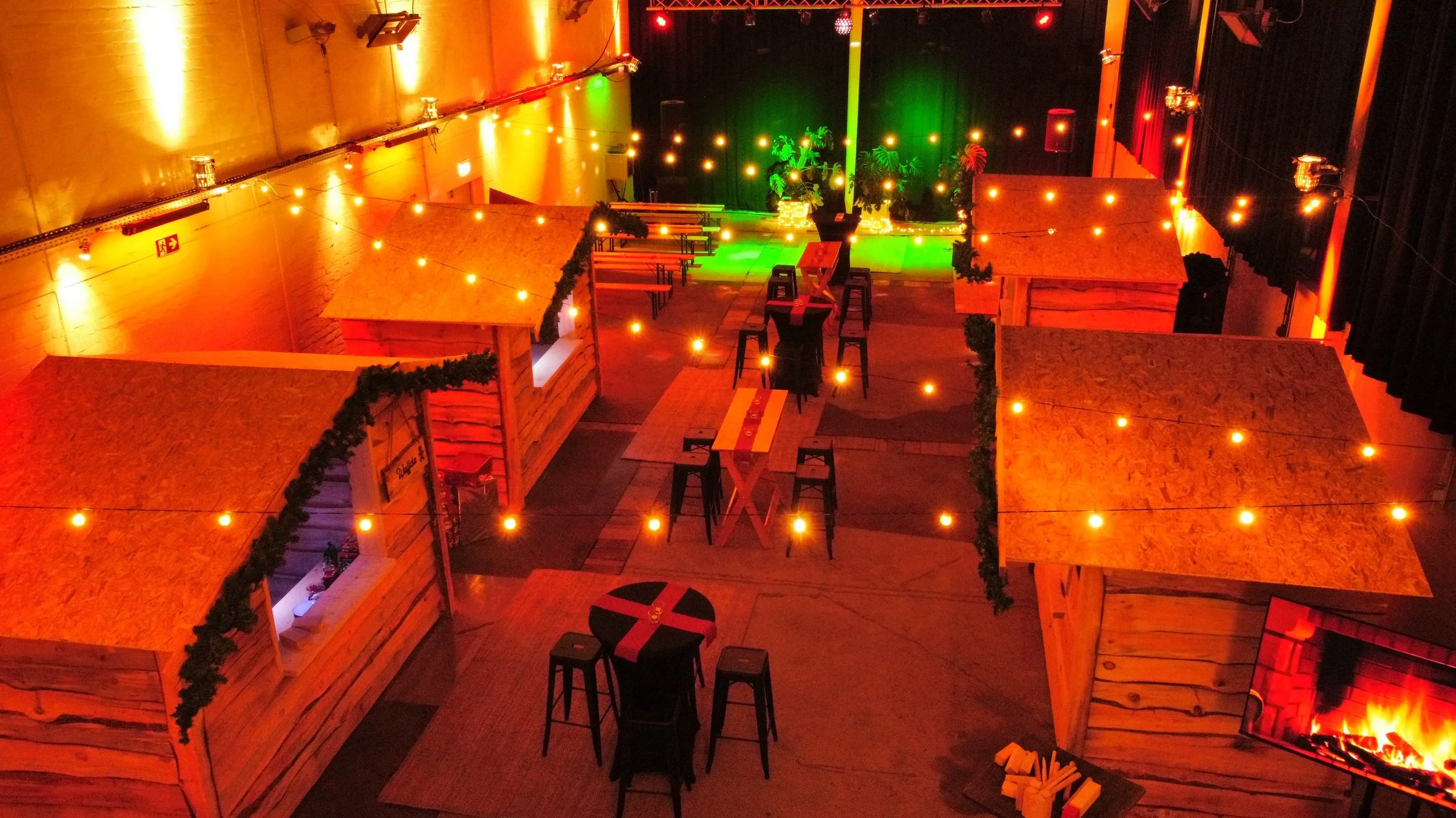 Eine stilvolle Bar- oder Lounge-Umgebung mit Holztischen und Bänken, dekorativen Lichterketten, Pflanzen und einer kleinen Bühne mit einer Green- und Rott-Lichtkunst, sowie einer Feuerstelle im unteren rechten Bereich.