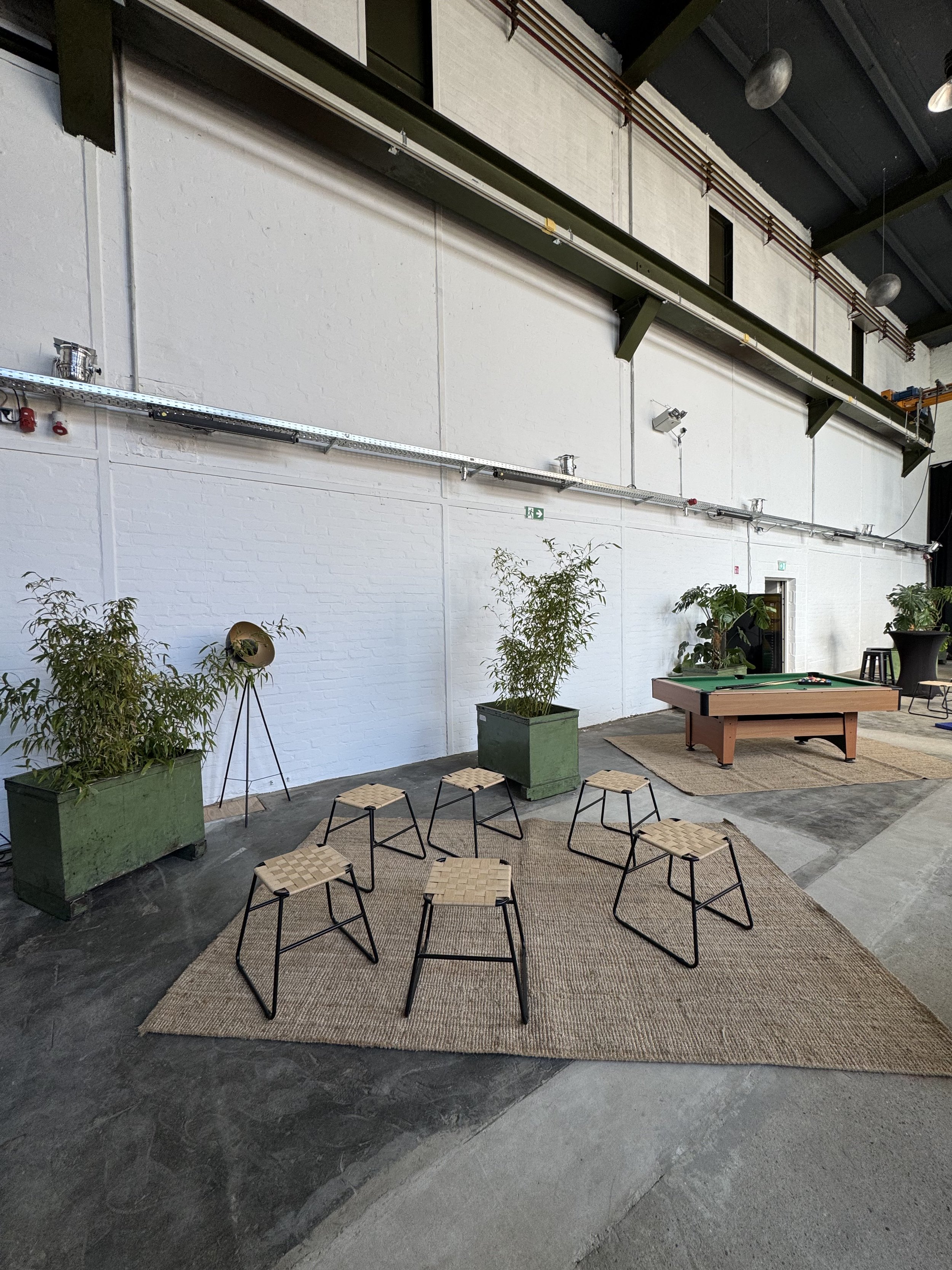 Innenansicht eines modernen, offenen Raumes mit weißen Wänden, grünen Pflanzen in großen Töpfen, einem Billardtisch, Stühlen, Teppichen und einer hohen Decke mit schwarzen Metallträgern und Beleuchtung. Es scheint ein Veranstaltungsraum oder Lounge zu sein.