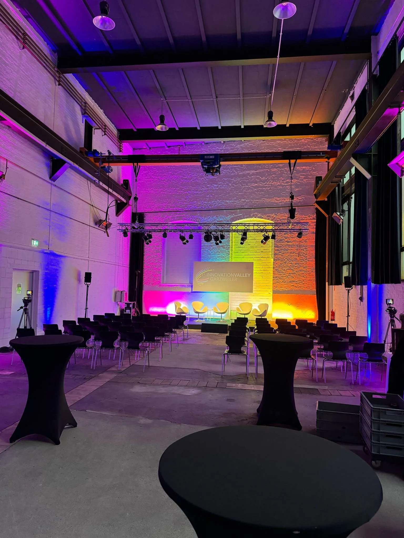 Live-Event-Bühne mit Stühlen, Stehtischen und bunten Lichteffekten, die die Wände beleuchten, im Raum der Innovations Valley Garzweiler.