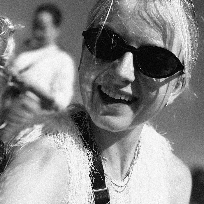 A woman wearing sunglasses and a sleeveless top smiling outdoors with another woman blurred in the background.