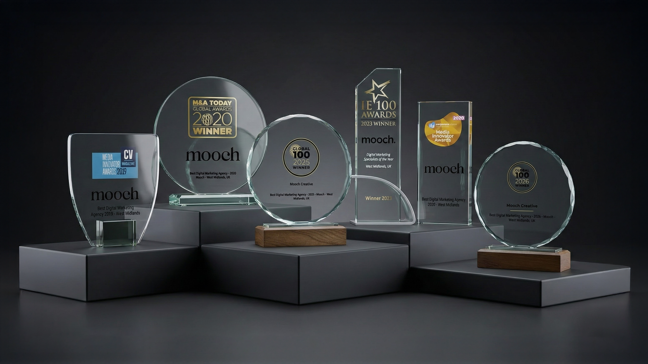 Collection of six glass award trophies with engraved text, displayed on gray platforms against a dark background.