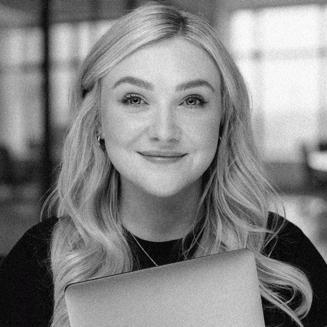 A woman with long wavy blonde hair smiling, wearing makeup and a nose ring, in a dimly lit indoor setting.