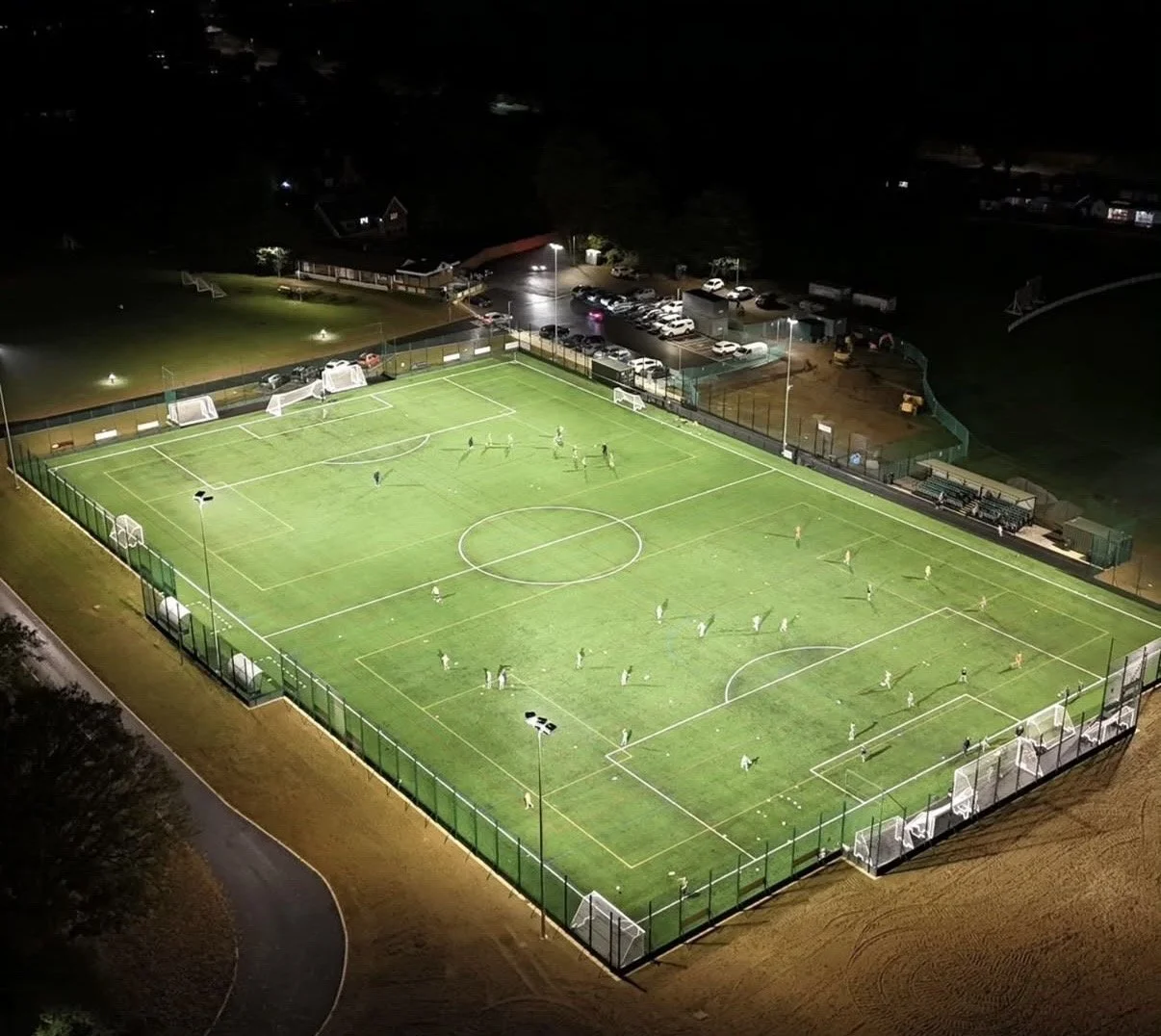 Aerial night view of a well-lit soccer field with players practicing or playing, surrounded by a fence, with parking lot and some buildings visible in the background.