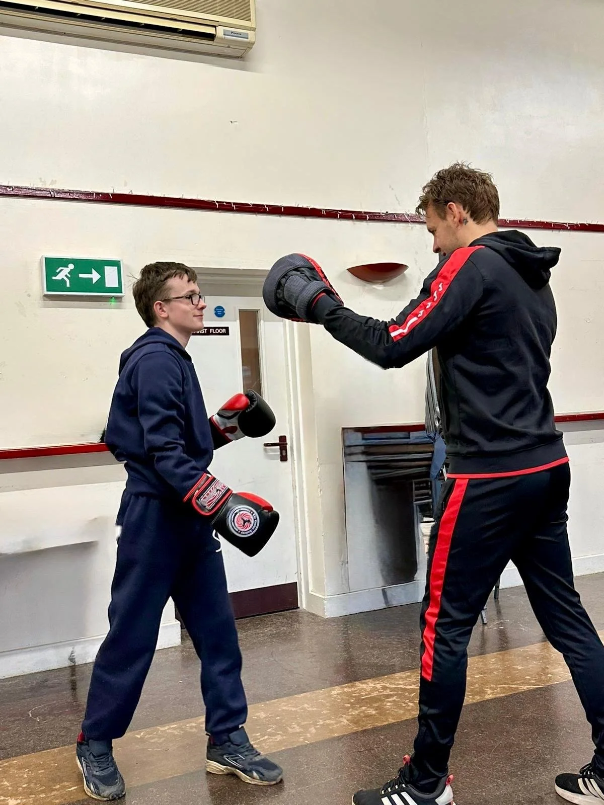 Two young men training boxing indoors, one is a teenager with glasses and the other appears to be an adult, both wearing sportswear and holding boxing gloves.