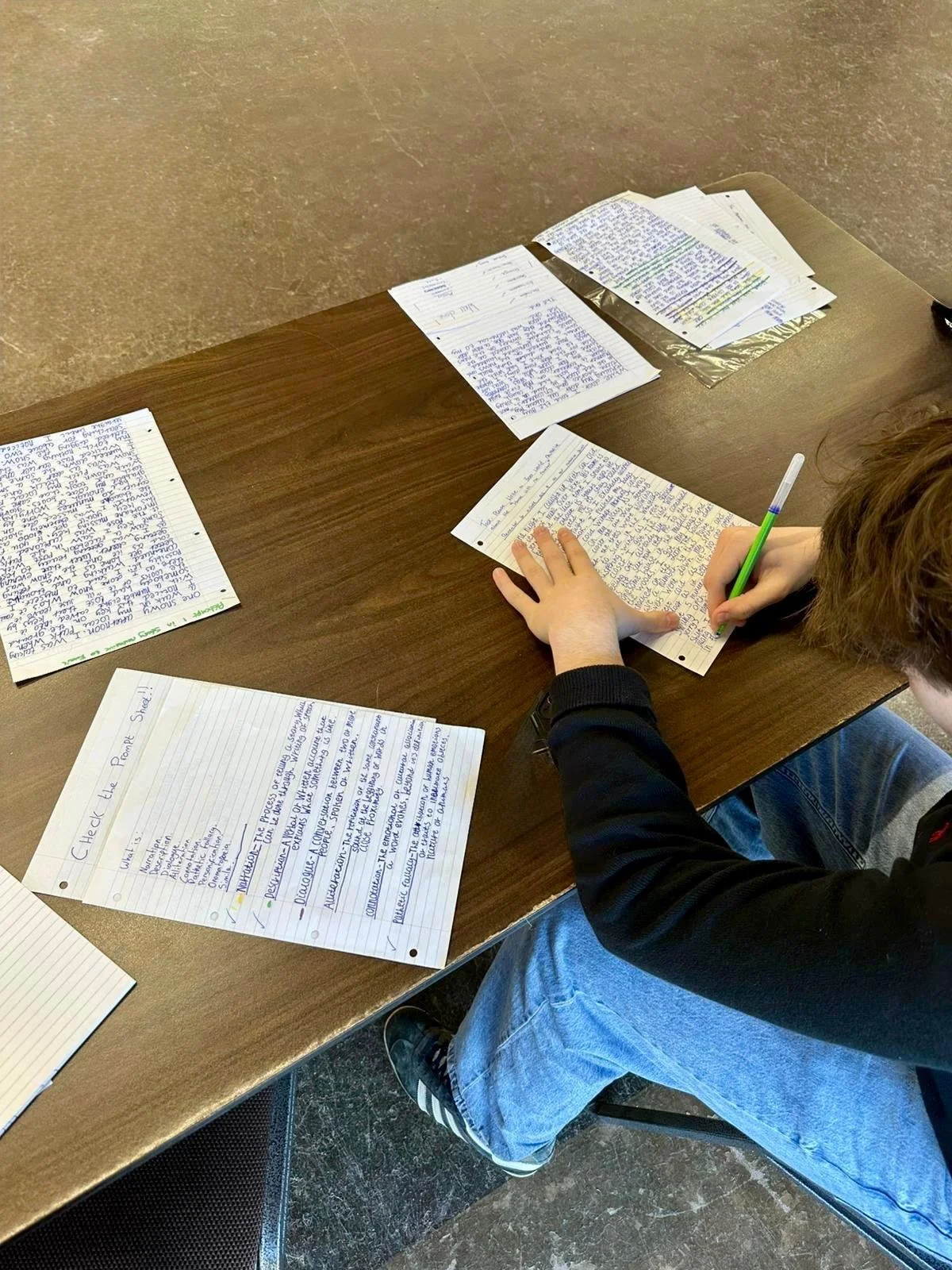 A person sitting at a wooden table writing on lined paper, with several other handwritten papers spread out on the table.