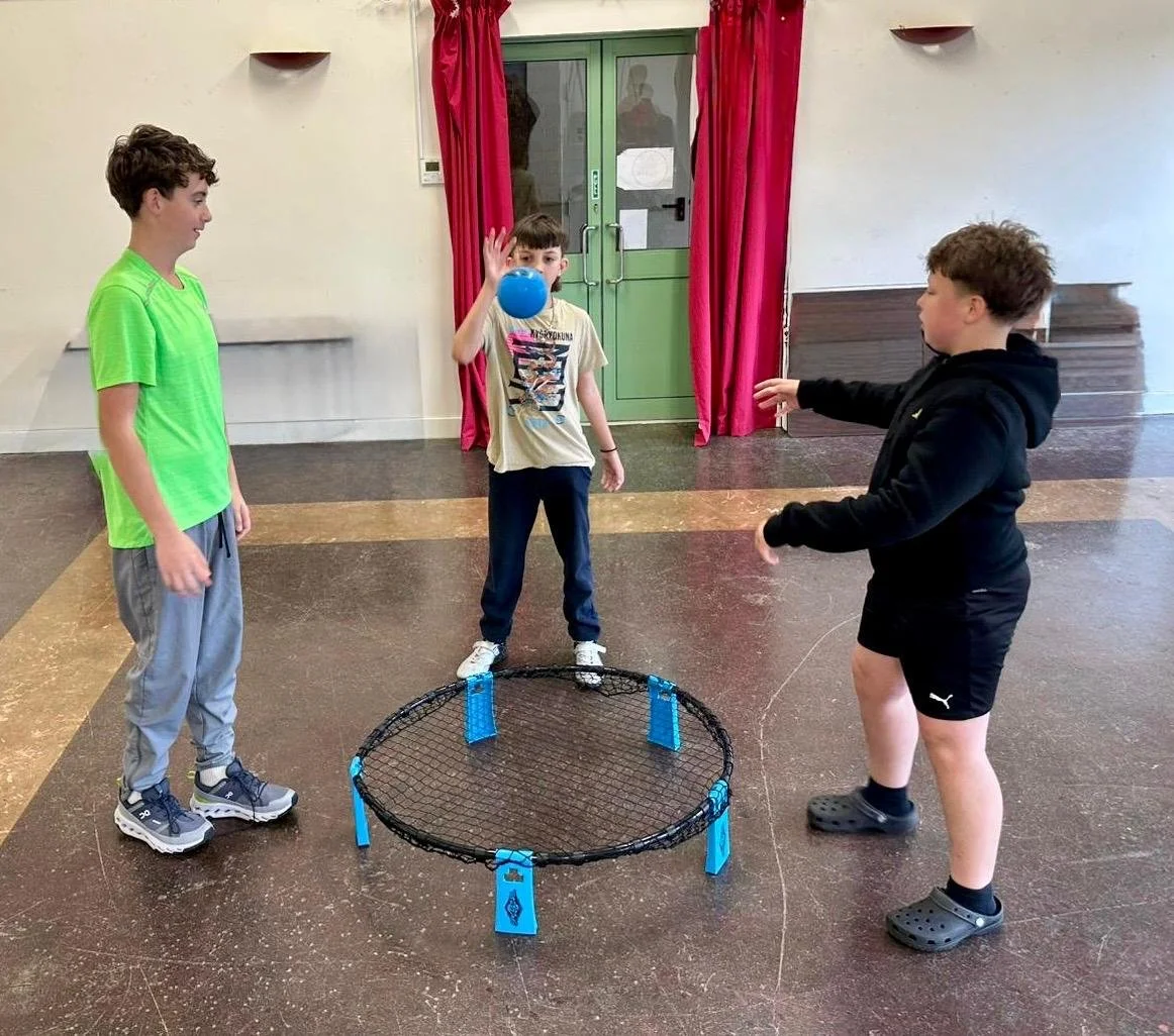Group of seven young women standing inside near a glass door, holding boxes of headphones, with an outdoor sports field visible through the glass.