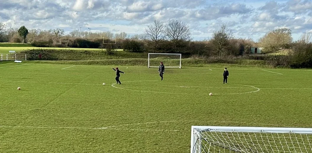 Three people playing soccer on a grassy field with goalposts, soccer balls, and trees in the background.