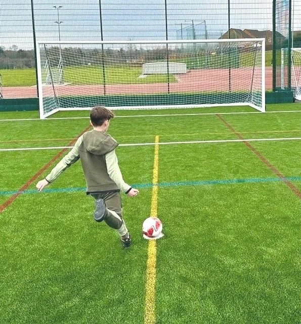 A young boy is kicking a soccer ball on a green artificial turf field, aiming at a soccer goal in front of him.
