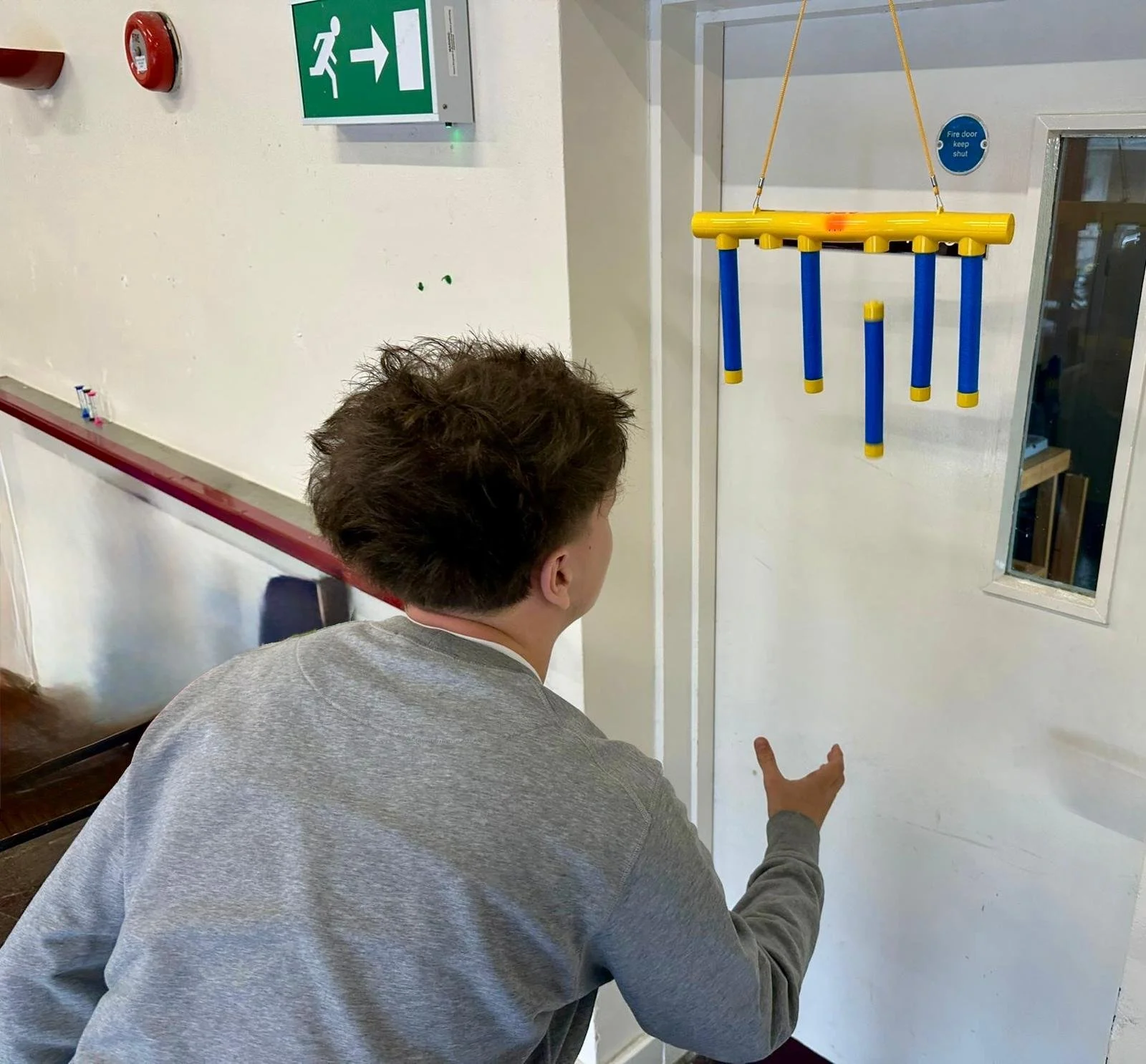 View of a nearly empty soccer stadium with red seats and the abbreviation "WEMBLEY" spelled out in seats. Several young men are sitting in the stands, some looking at their phones, with a view of the green field below.