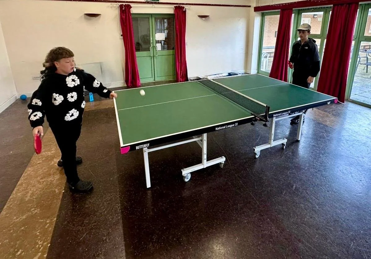 Two children playing table tennis indoors, with a girl on the left holding a paddle and a boy on the right standing by the table, ready to hit the ball.