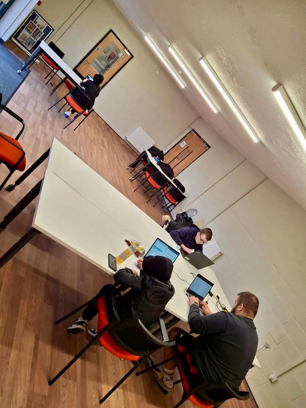 A room with a long white table, several people using laptops, and chairs with orange cushions. There are additional chairs and tables along the back wall, a door, windows, and fluorescent lighting on the ceiling.