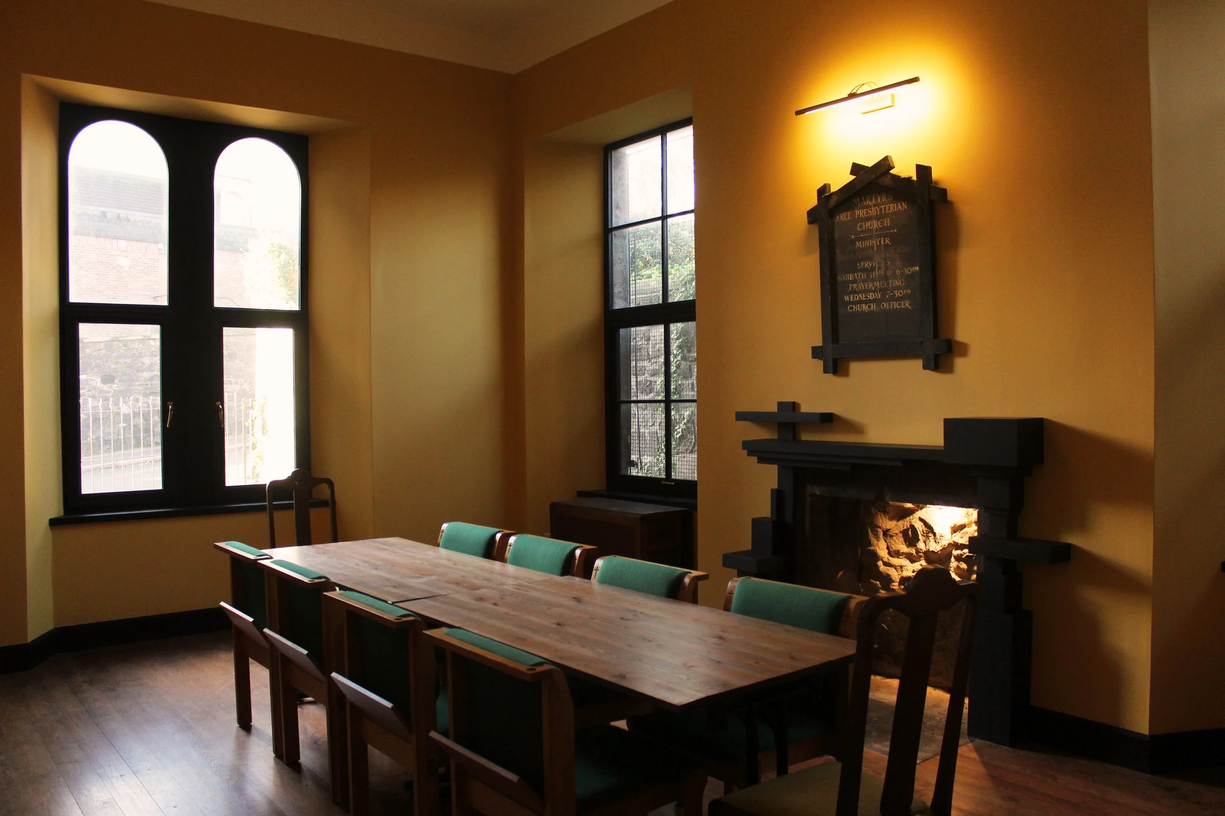 Interior of a room with a long wooden table, green chairs, a fireplace, large windows, and a plaque on the wall.