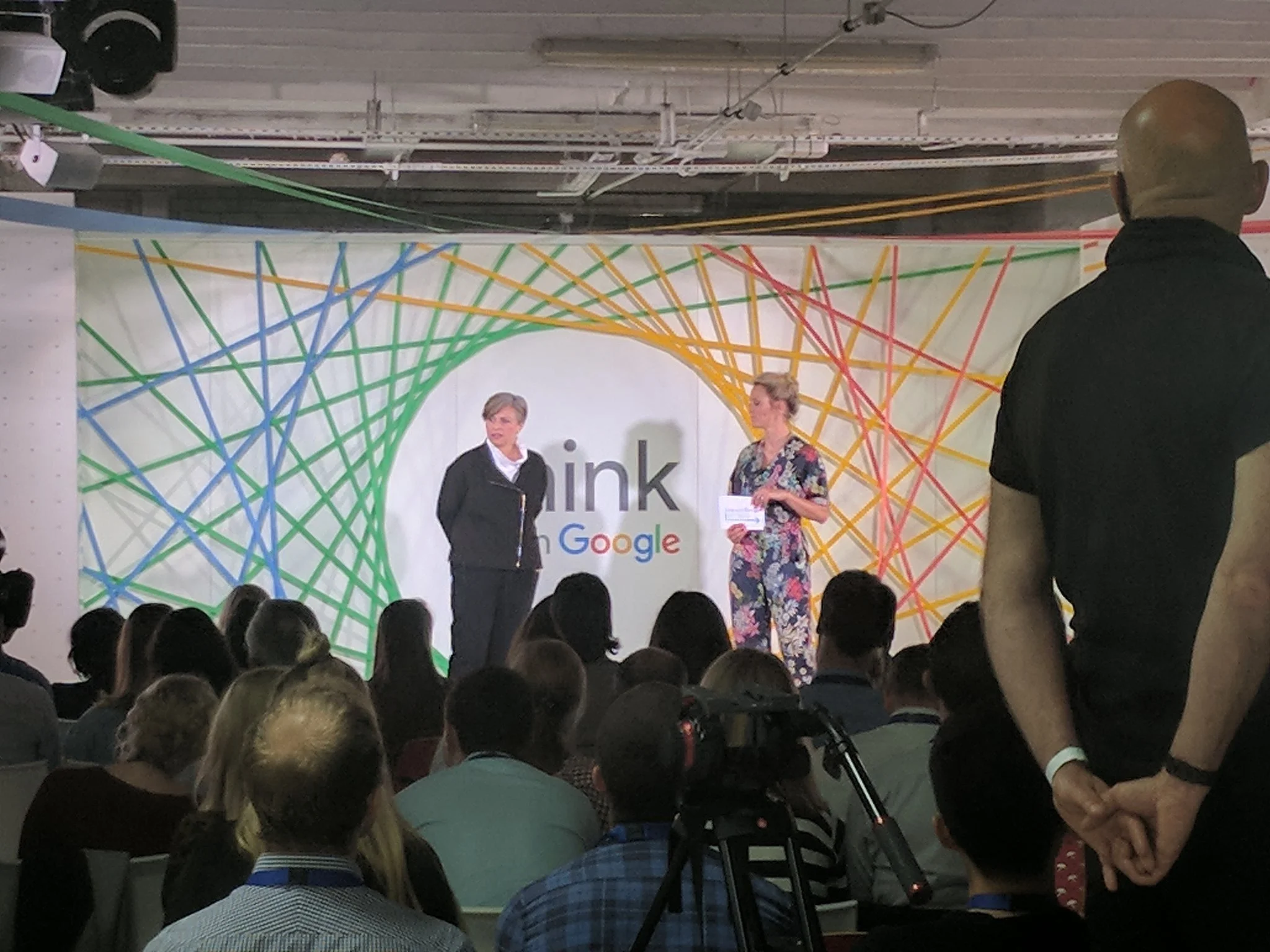 A woman who Tracey Follows is speaking on stage at a Think with Google event, with a large colorful background design featuring interconnected lines and the Google logo in front of a large audience 