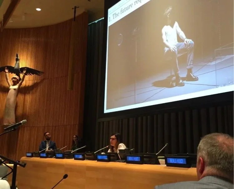 A woman (Tracey Follows) speaking at at teh UN HQ with a large screen behind her displaying a slide, and a sculpture of a person with arms outstretched on the left side of the stage.