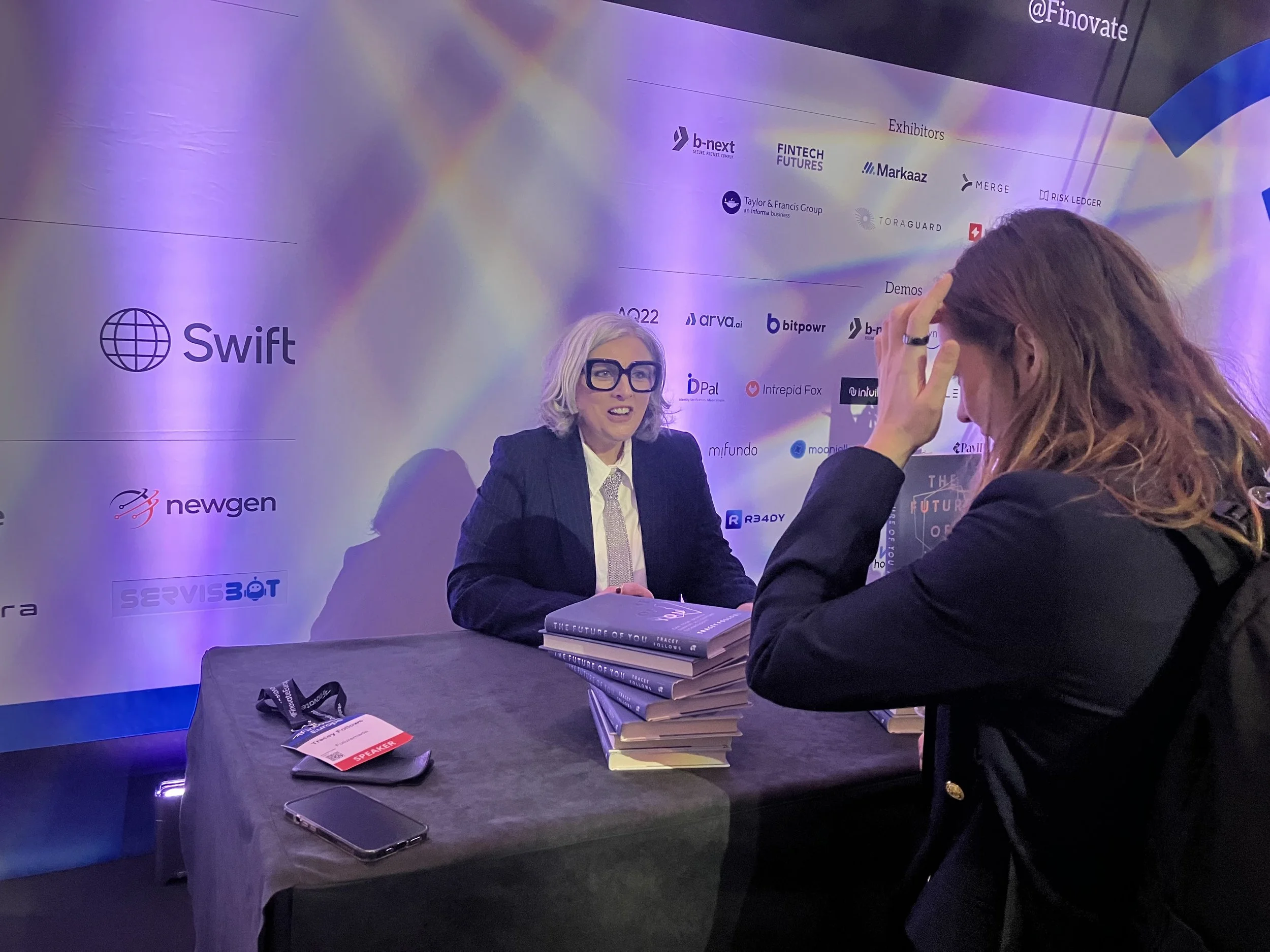 Tracey Follows, a woman with silver hair, glasses, and a dark blazer sits at a table stacked with books, talking to a woman with long brown hair and a black blazer in a conference or interview setting with a backdrop displaying various sponsor logos.