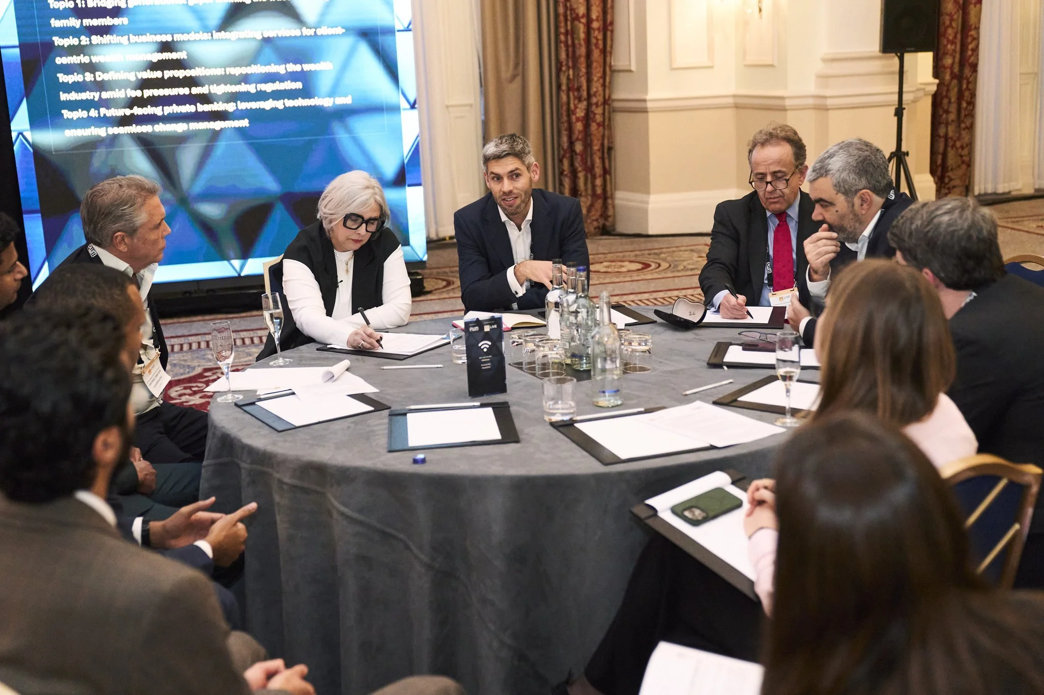 Tracey Follows with a group of diverse professionals engaged in a discussion around a round conference table iat an FT Wealth Summit event n a formal setting with a large screen displaying presentation topics in the background.