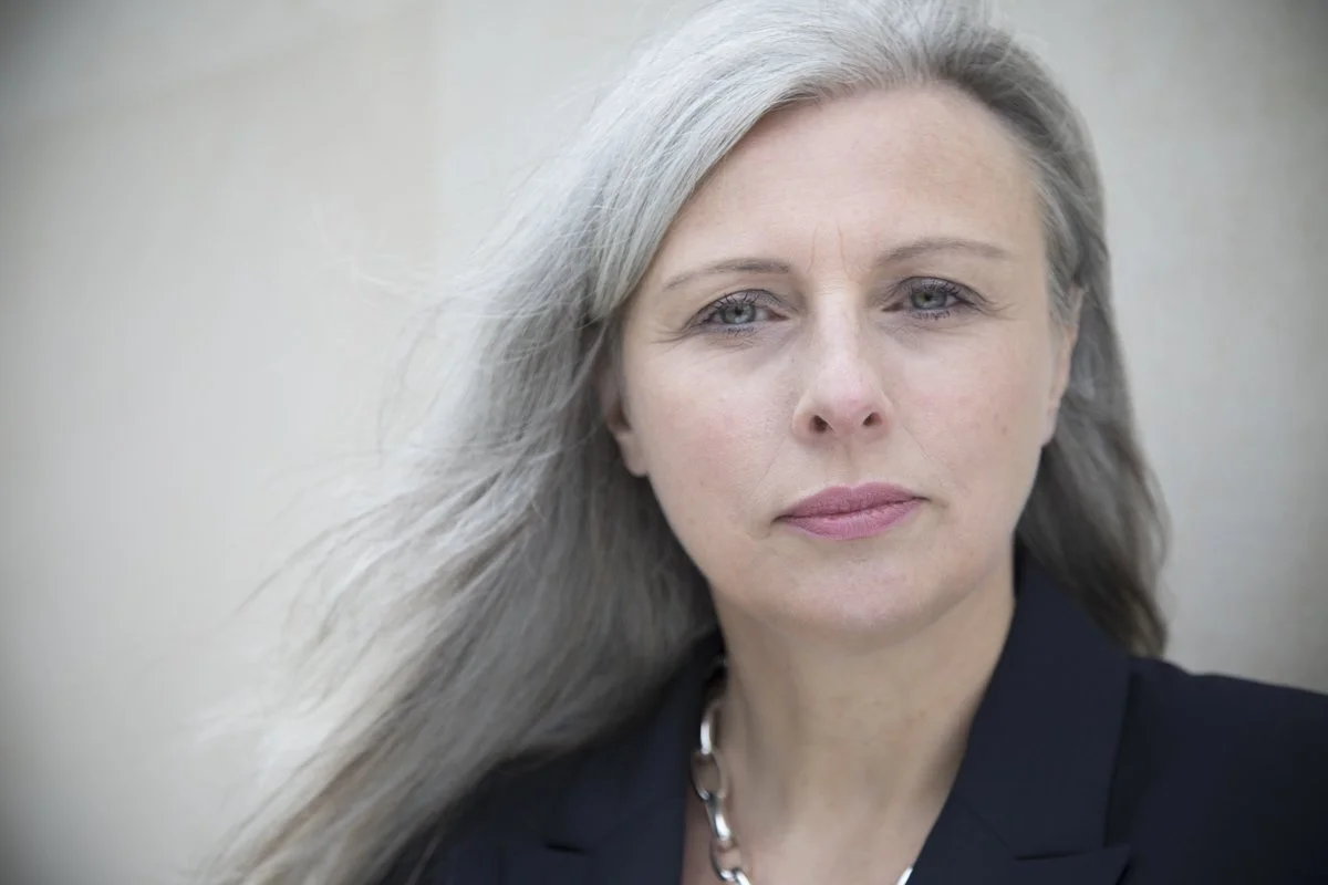 Tracey Follows with long silver  hair and blue eyes wearing a black blazer and a silver necklace.
