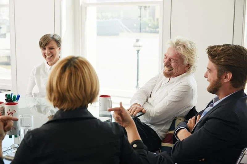 Tracey Follows and Sir Richard Branson plus two other people in a business meeting or discussion, sitting around a glass table with mugs and pens, smiling and engaging in conversation, in a bright room with large windows.