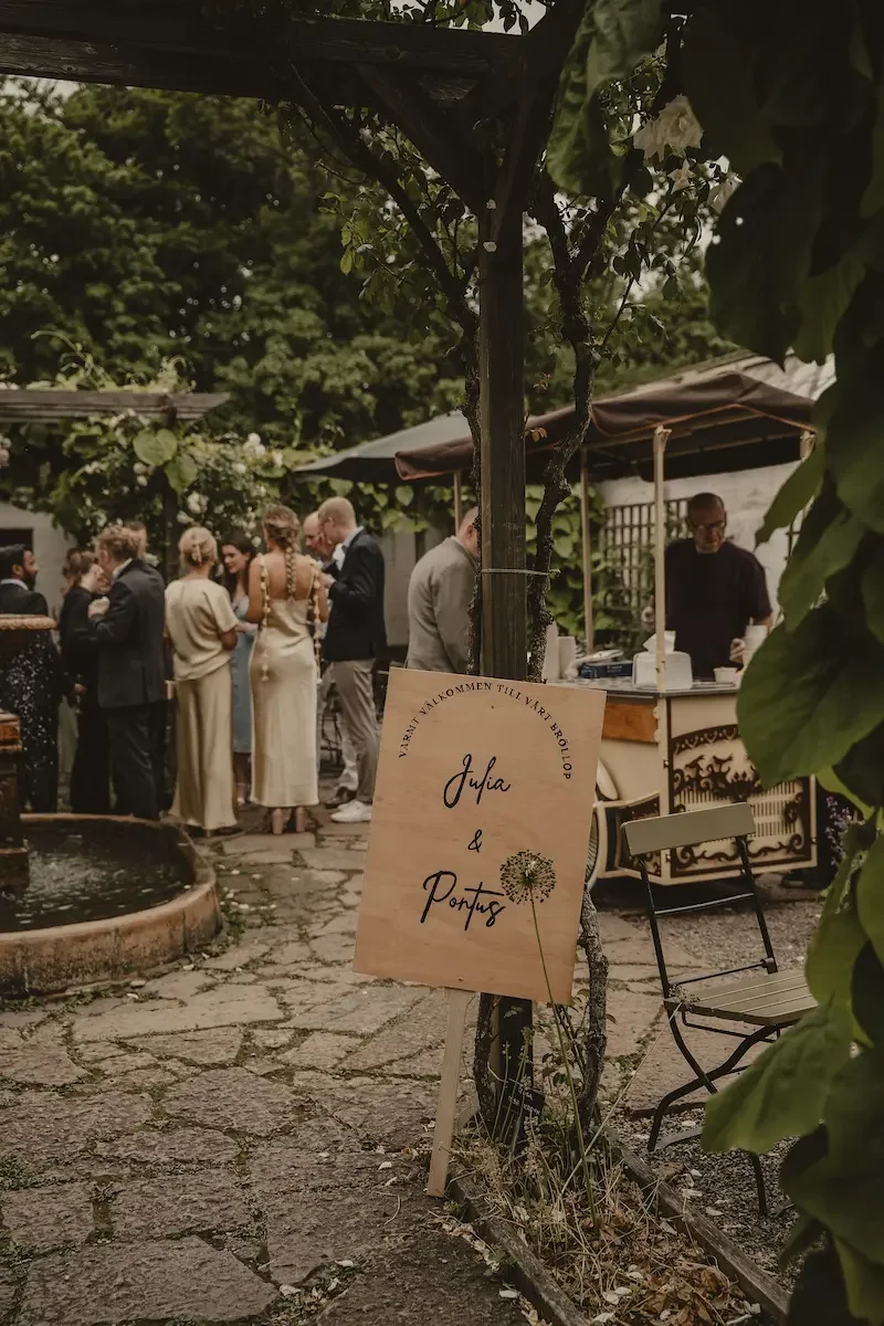 Träskylt med brudparets namn som välkomnar gäster till bröllopsmingel i trädgårdsmiljö på Katrinetorp Landeri.