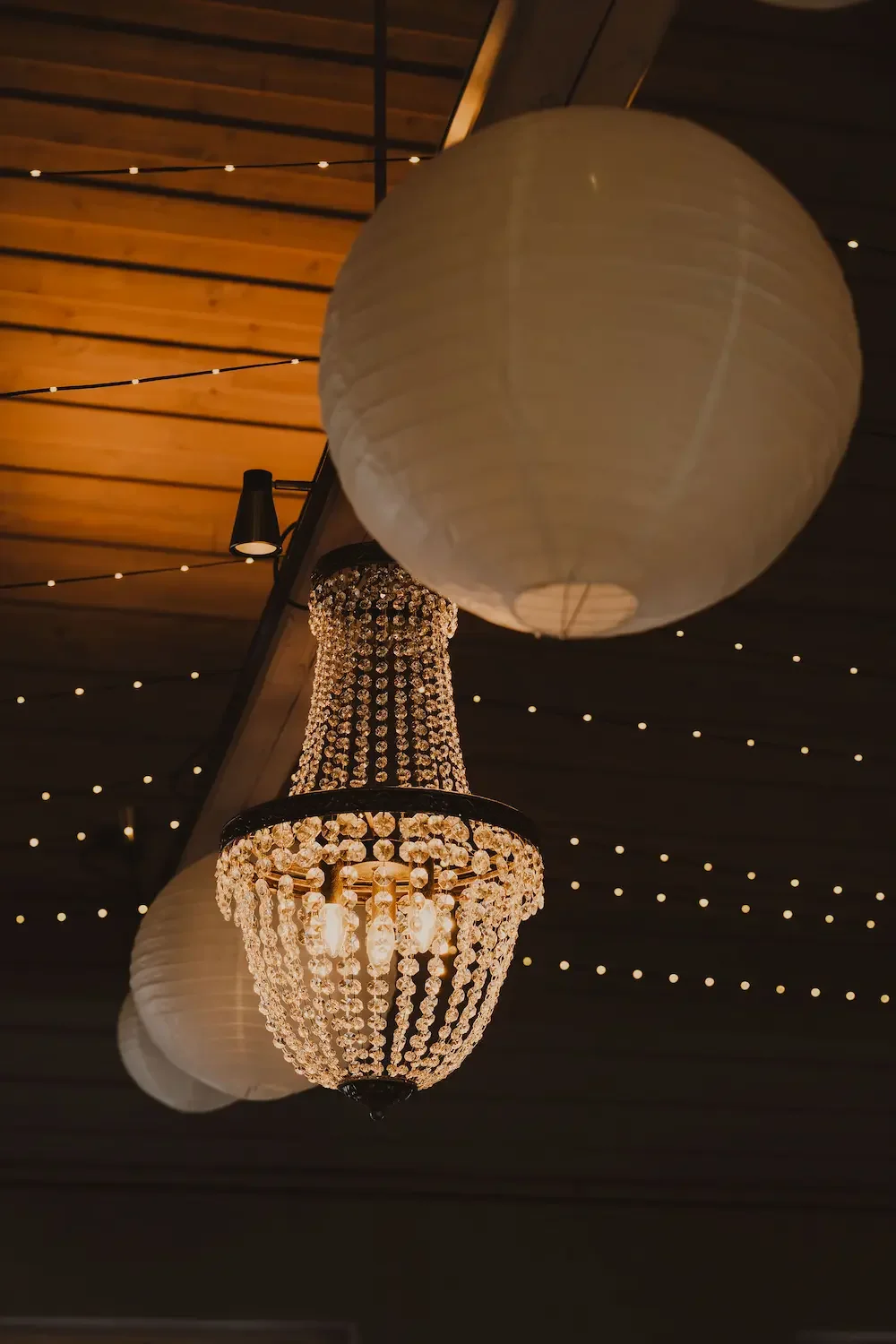 Stämningsfull närbild på bröllopsdekorationer i taket med takkrona, ljusslingor och risbollar. 