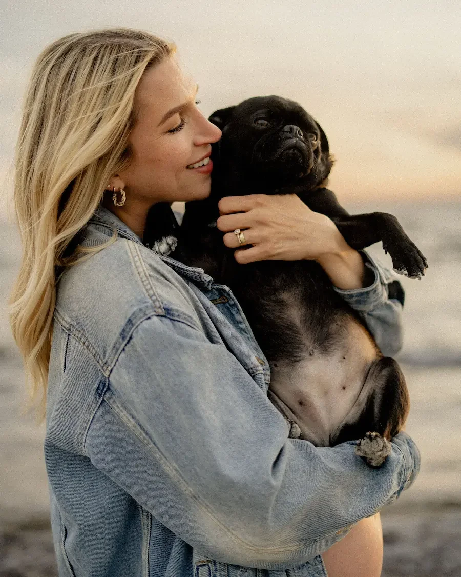 Gravid kvinna i jeansjacka som kramar sin hund på en strand i Skåne under en gravidfotografering.