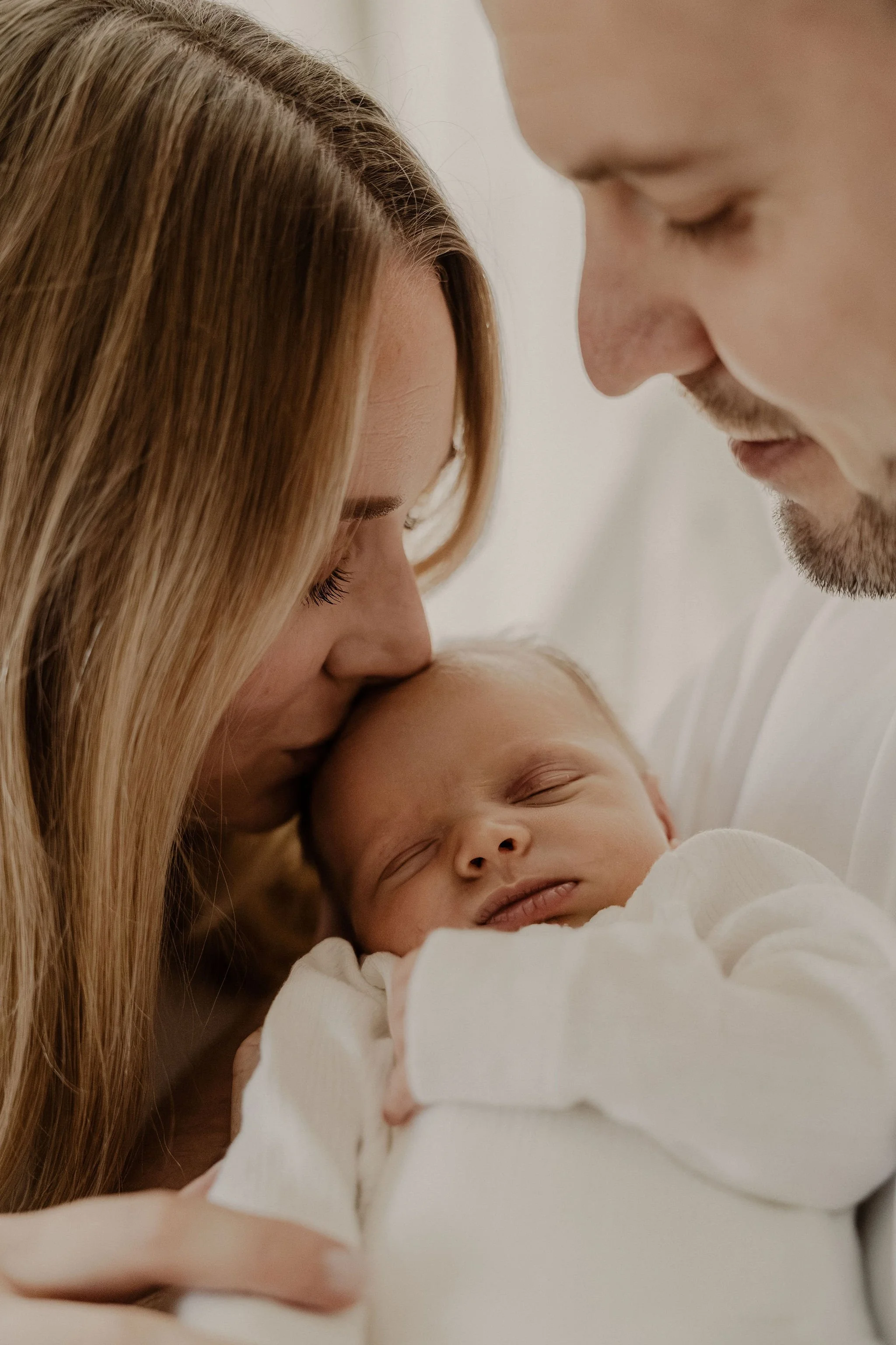 En nära och känslofull bild från en nyföddfotografering, där två föräldrar håller sin bebis mellan sig. Mamman pussar bebisen på huvudet och pappan tittar kärleksfullt.
