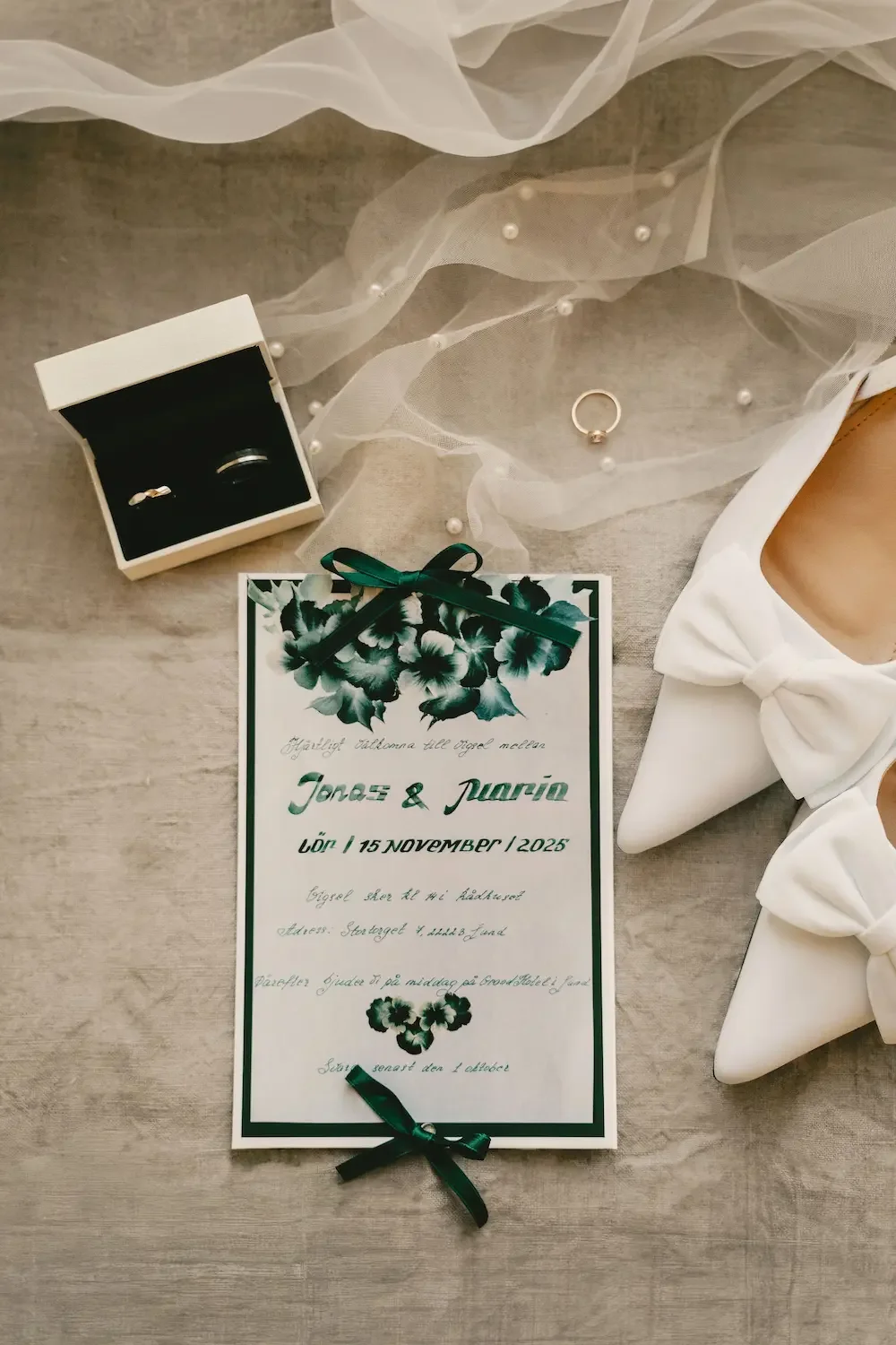 Flatlay på bröllopsdetaljer med inbjudan i mitten omgiven av vigselringar, slöja och klacksko, modern och editorial stil.