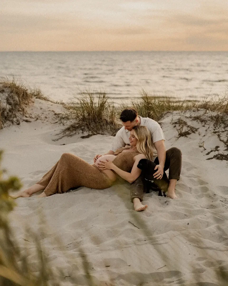 Ett par med en hund som myser bland sanddynorna i solnedgången på en strand i Skåne. De tittar ner på kvinnans gravida mage.