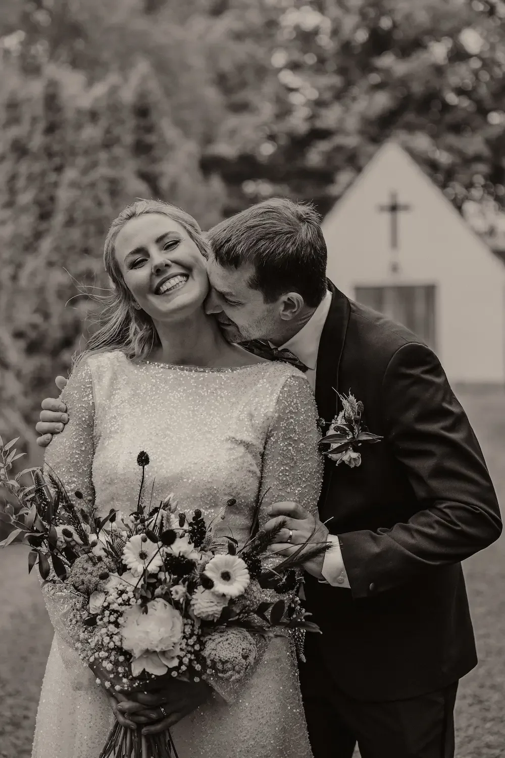 Intim svartvit bild utanför kyrka i Skåne där brudgummen kysser bruden på halsen medan hon skrattar. 