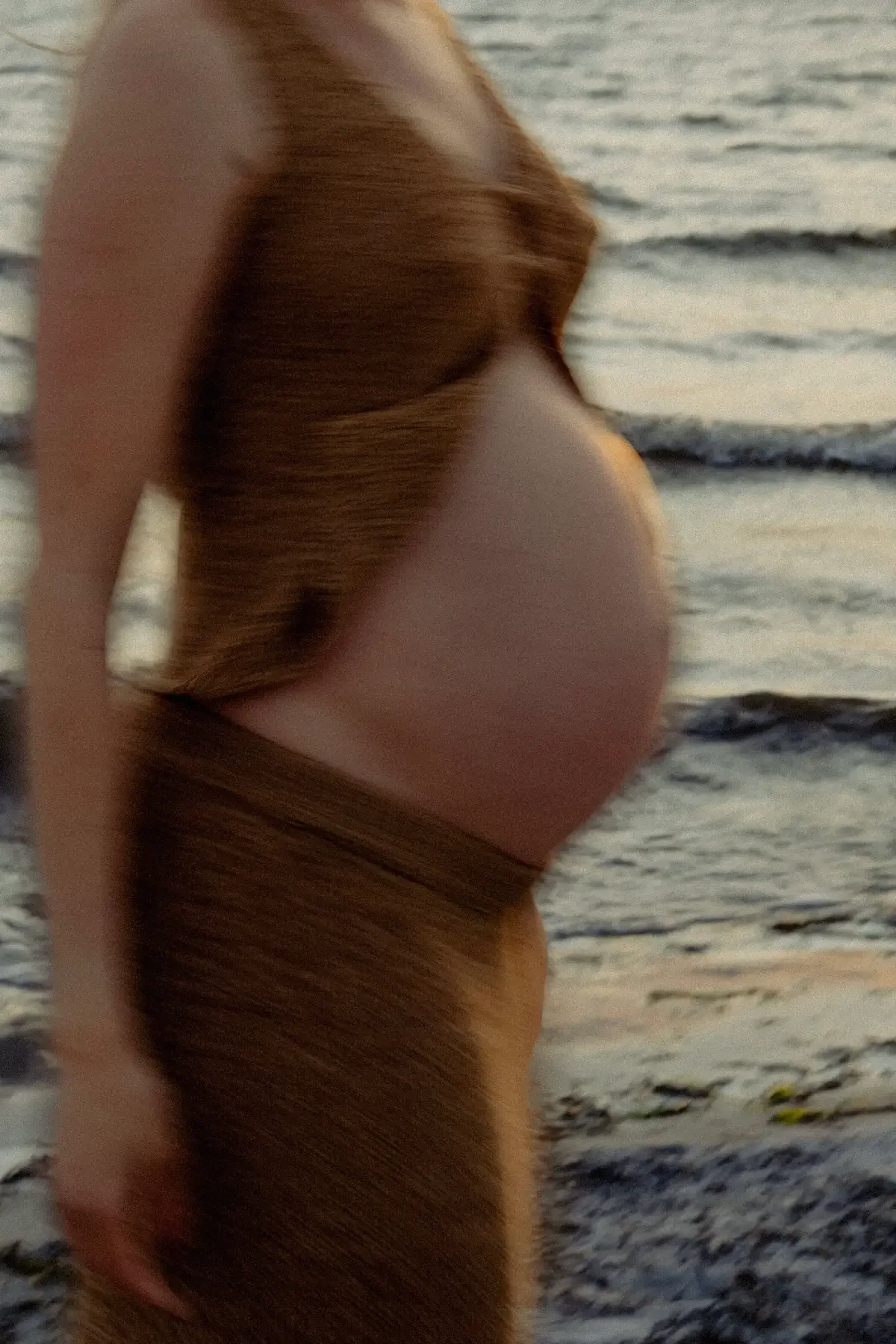 En gravid kvinna i jordnära kläder som går på en strand i solnedgången.