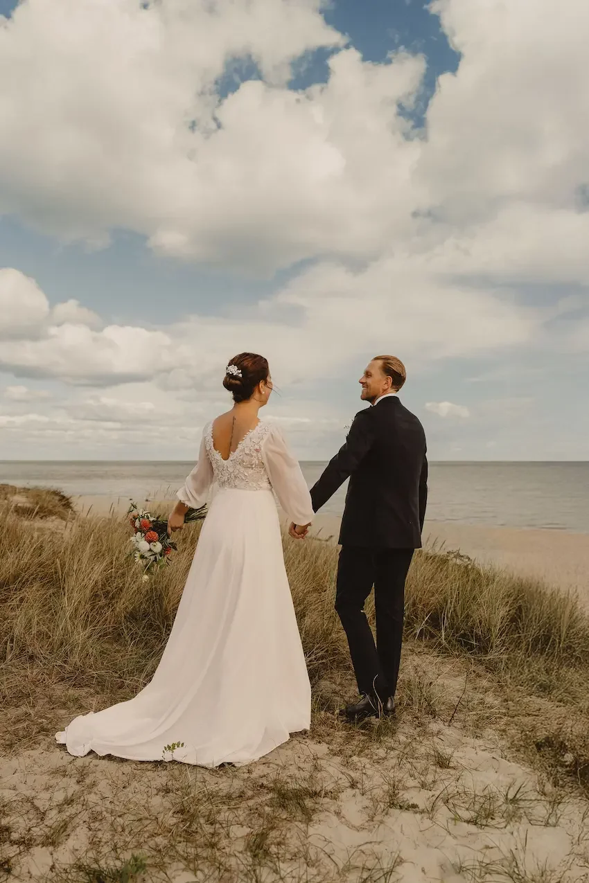 Brudpar går längs en strand på Österlen i Skåne med havet i bakgrunden.