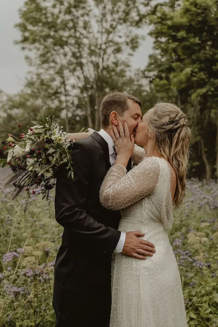 Brudpar med eleganta kläder och glittrig brudklänning kysser varandra vid blomsteräng i Skåne.
