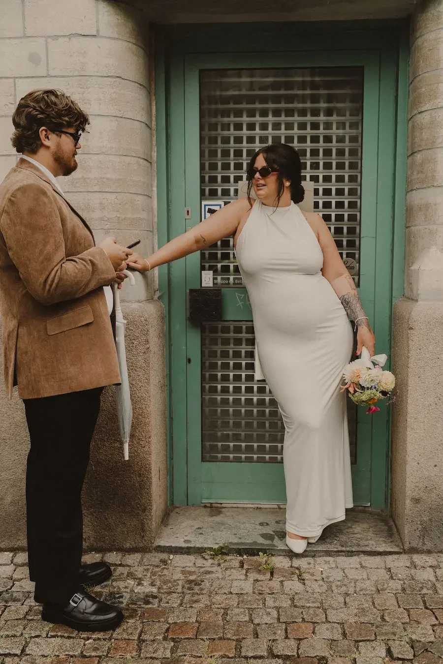 Brud som står i snygg pose och pratar med brudgummen under ett litet tak för att skydda sig från regnet i Göteborg medan de väntar på taxi.