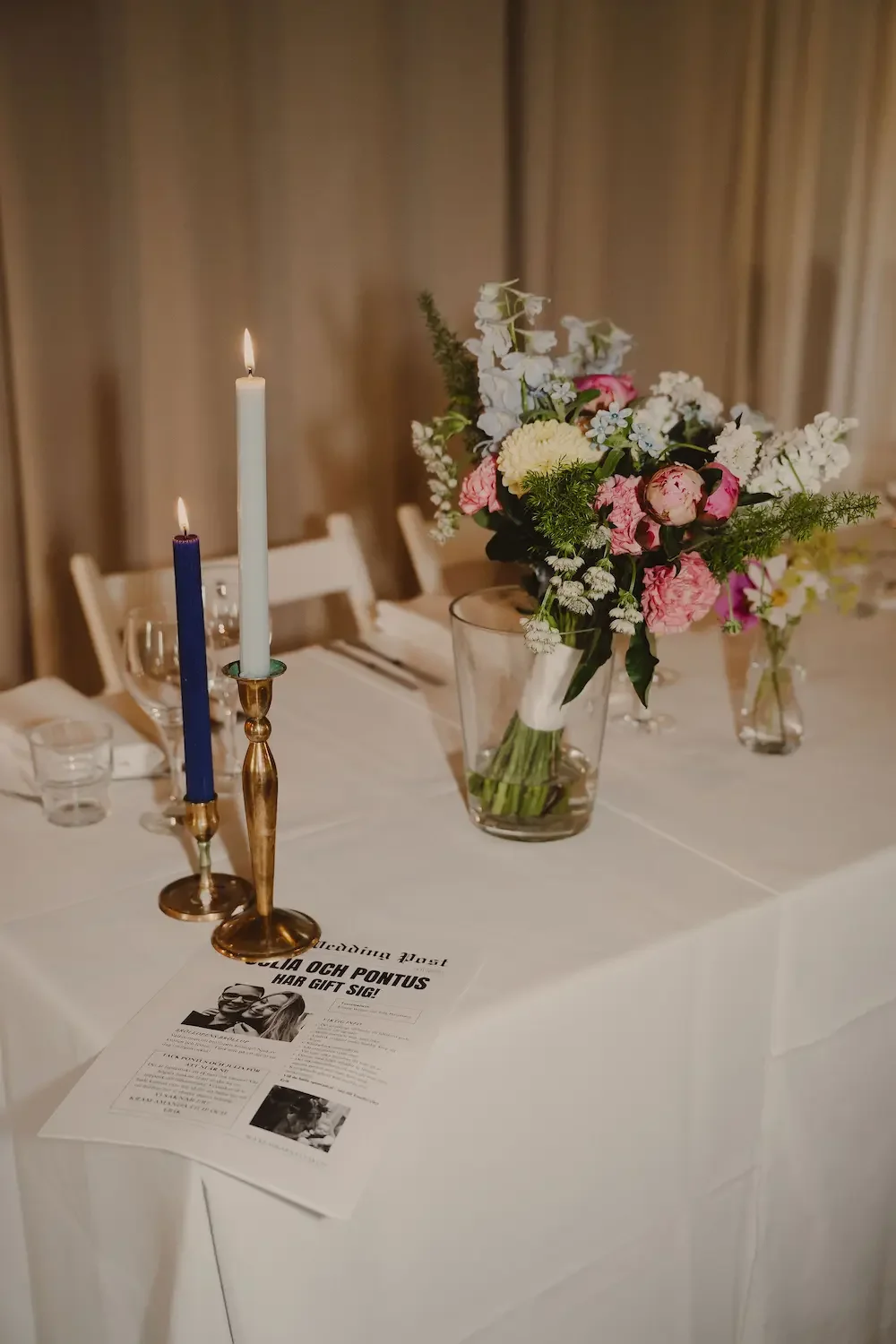 Detaljbild med färgglada ljus, brudbukett och en utskriven tidningsartikel om brudparet på ett dukat bord inför bröllopsmiddag.