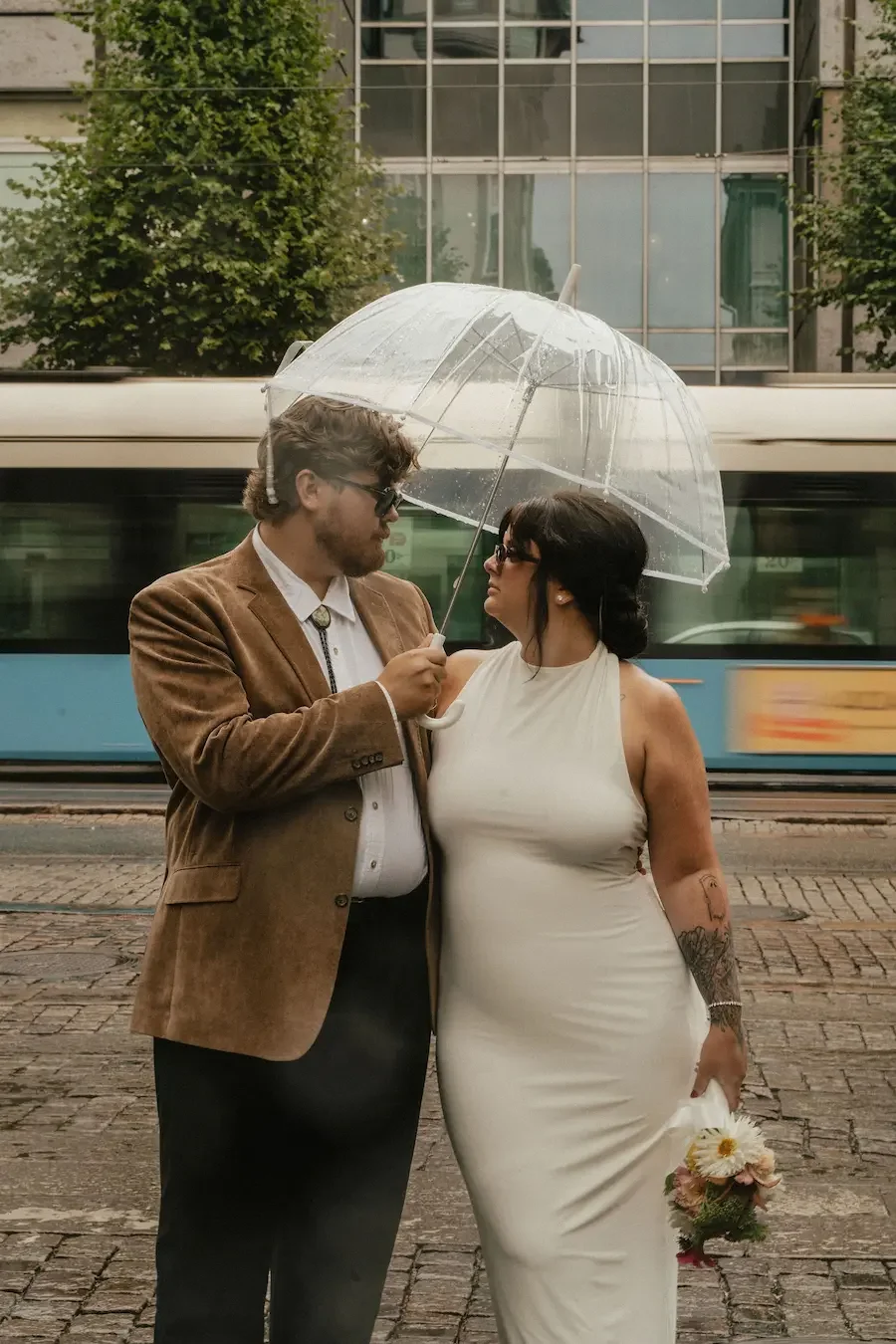 Brudpar som står på gatan i Göteborg under ett paraply i regnet, med en spårvagn som passerar i bakgrunden. Bilden fångar känslan av ett coolt, urbant stadsbröllop med modern, editorial känsla.
