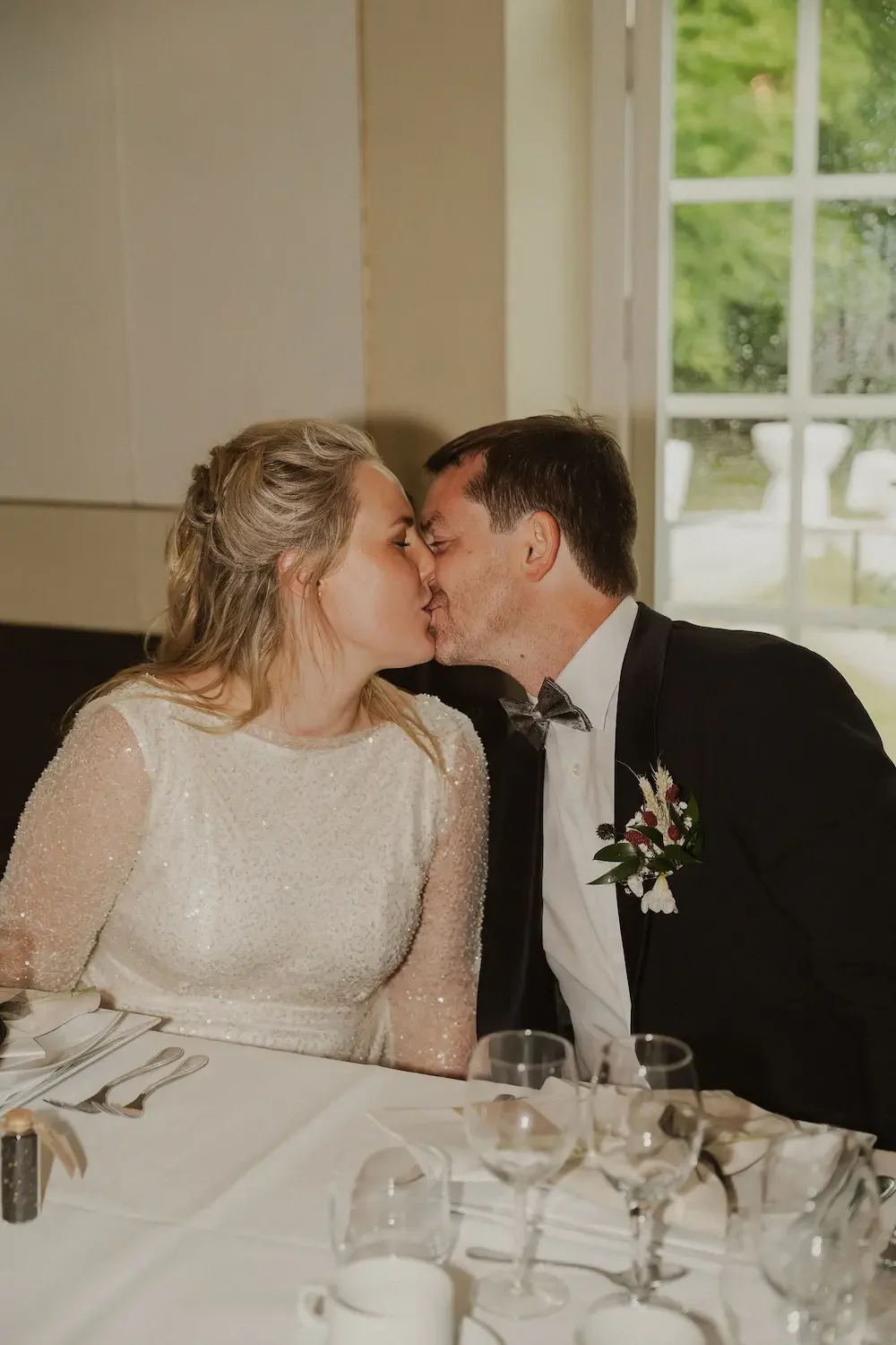Elegant brudpar kysser varandra vid middagsbordet på sin bröllopsfest i Skåne. 