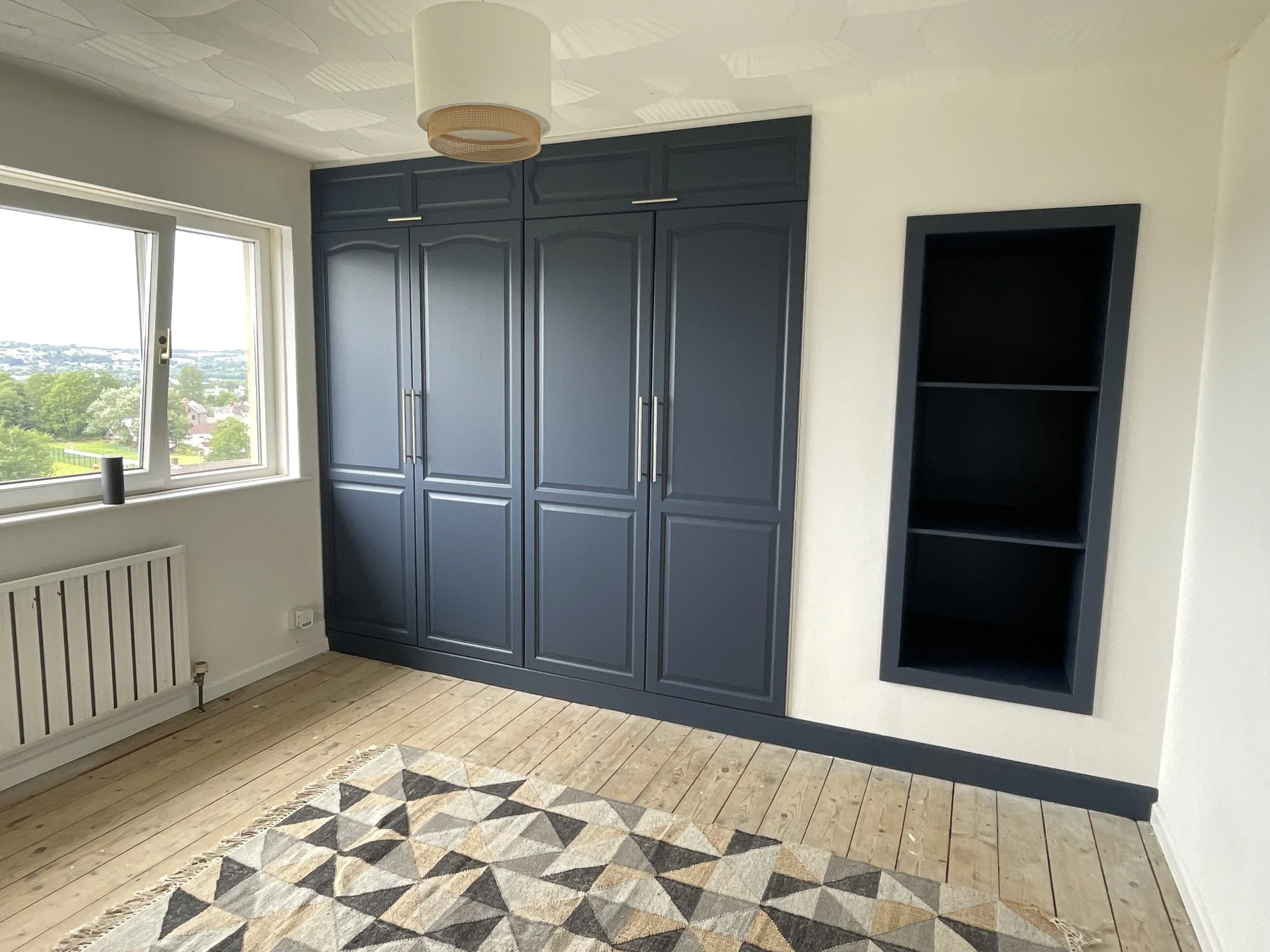 Room with a window showing a view of trees and buildings, a large dark blue closet with silver handles, and a black open shelf. The floor is wooden with a geometric patterned rug.