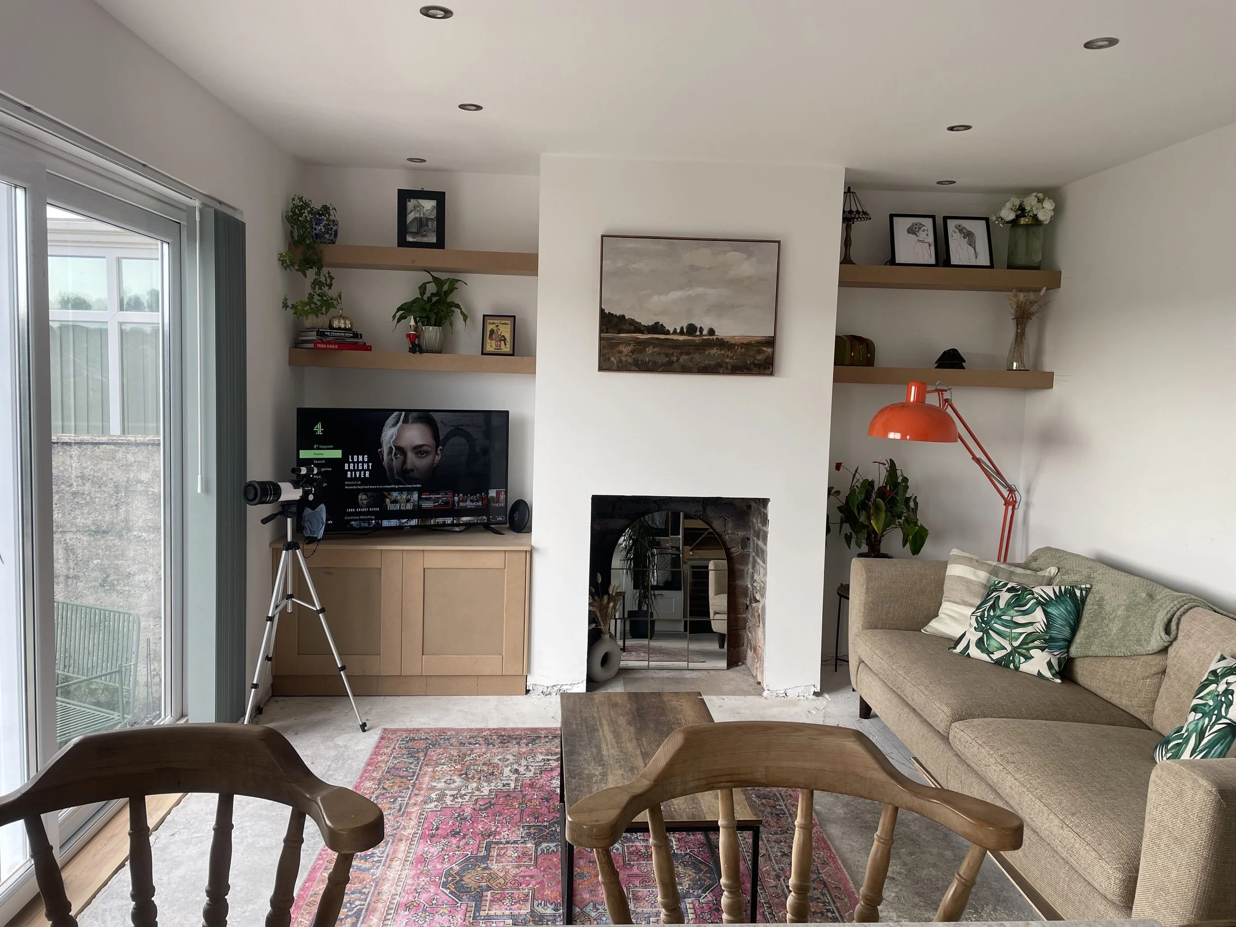 Living room with a beige sofa decorated with tropical leaf pillows, a wooden coffee table, a fireplace, a television with a telescope beside it, and floating shelves with plants and framed pictures. There is a sliding glass door on the left leading o