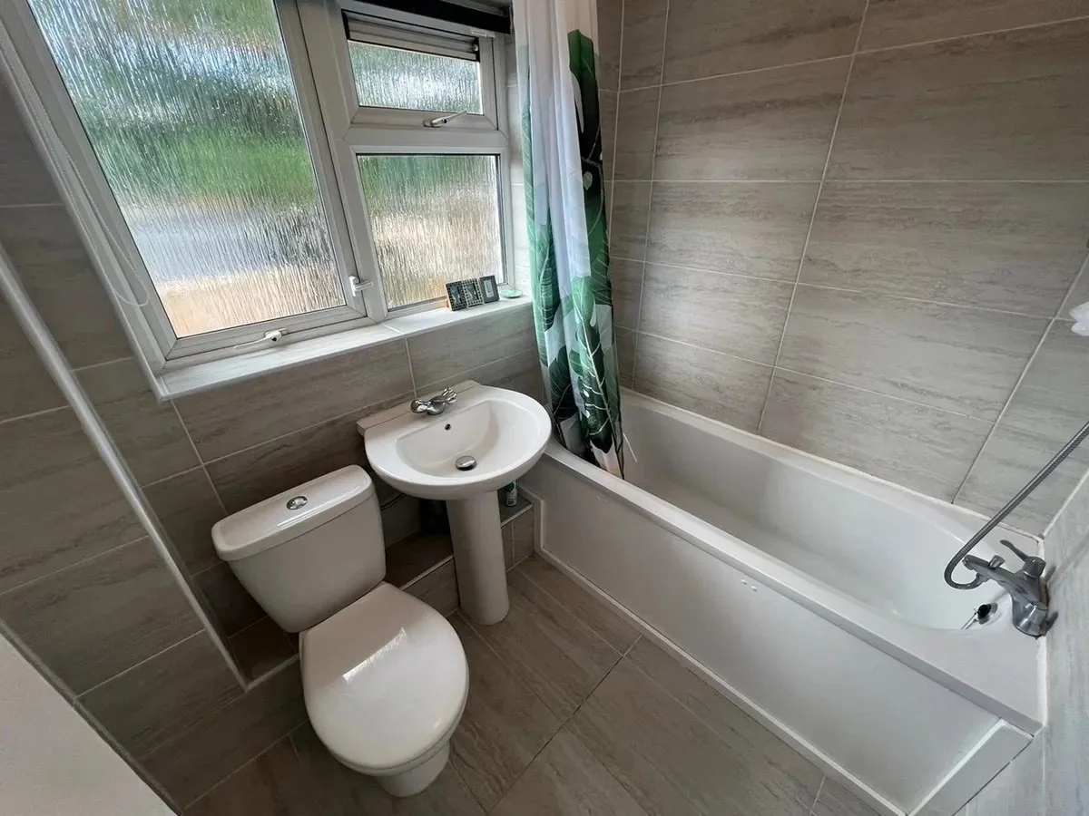 Bathroom with a toilet, small sink, bathtub, showerhead, and frosted window with a green and white shower curtain.