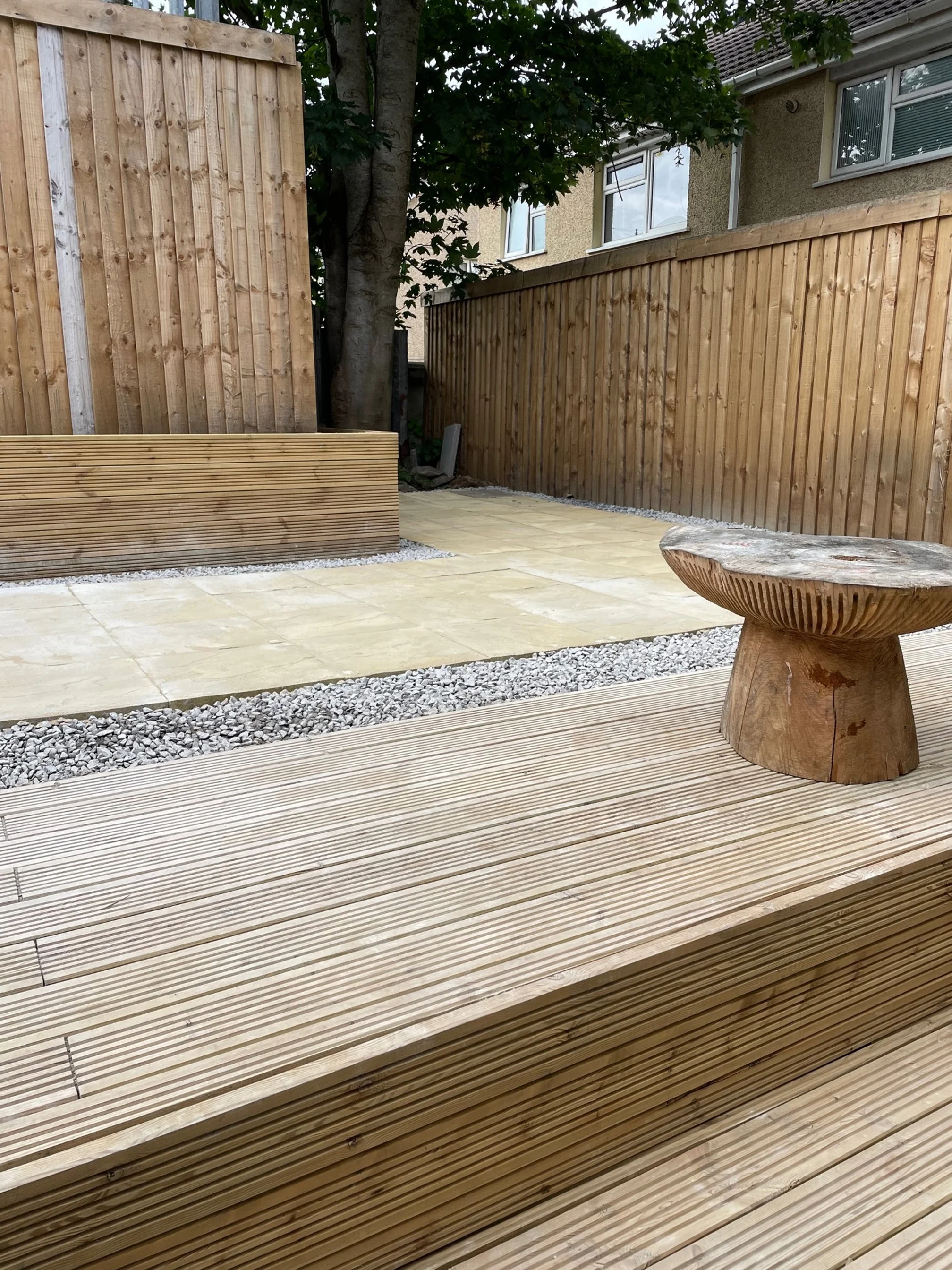 A backyard patio with wooden decking, a round wooden sculpture, a stone-paved area, a gravel strip, a wooden fence, and a tree with green leaves.