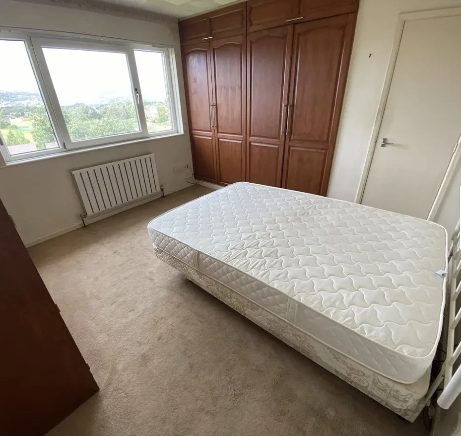 A bedroom with a bare mattress on the bed frame, wooden built-in closets, a large window showing a view of green trees and a cloudy sky, beige carpet, and a white door.