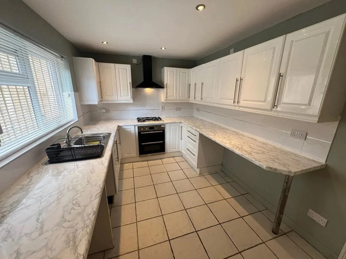 A clean, modern kitchen with white cabinets, marble countertops, a black oven and stove, a black range hood, a sink with a dish rack, and a window with blinds.