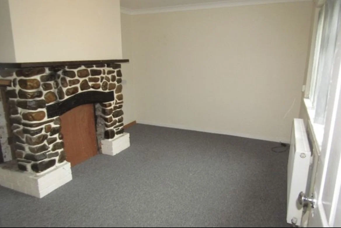 Empty room with gray carpet, a stone fireplace, beige walls, a large window with white curtains, and a white radiator on the wall.