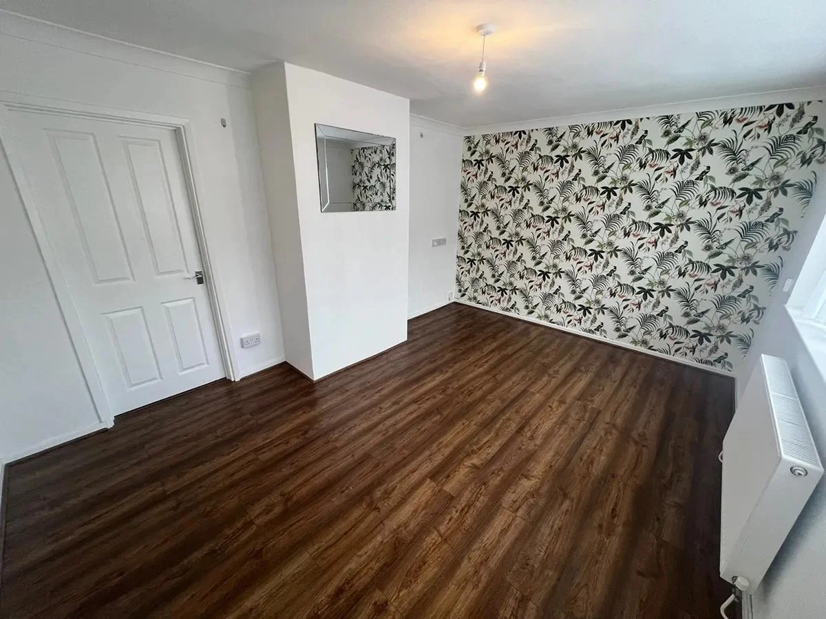 Empty living room with dark wood flooring, a white door, a wall with floral wallpaper, a mirror, and a radiator.