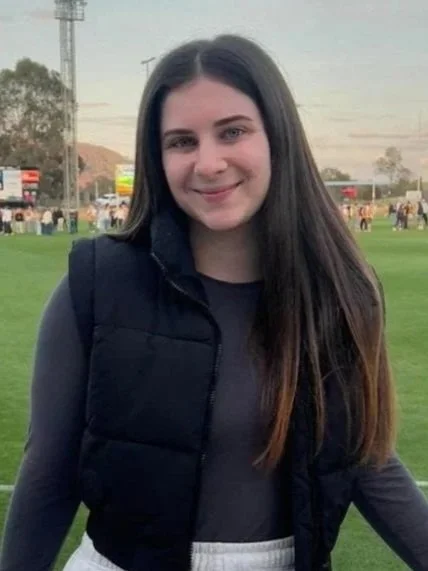 Person with long dark hair wearing a black vest and light gray pants standing in front of a sports field.