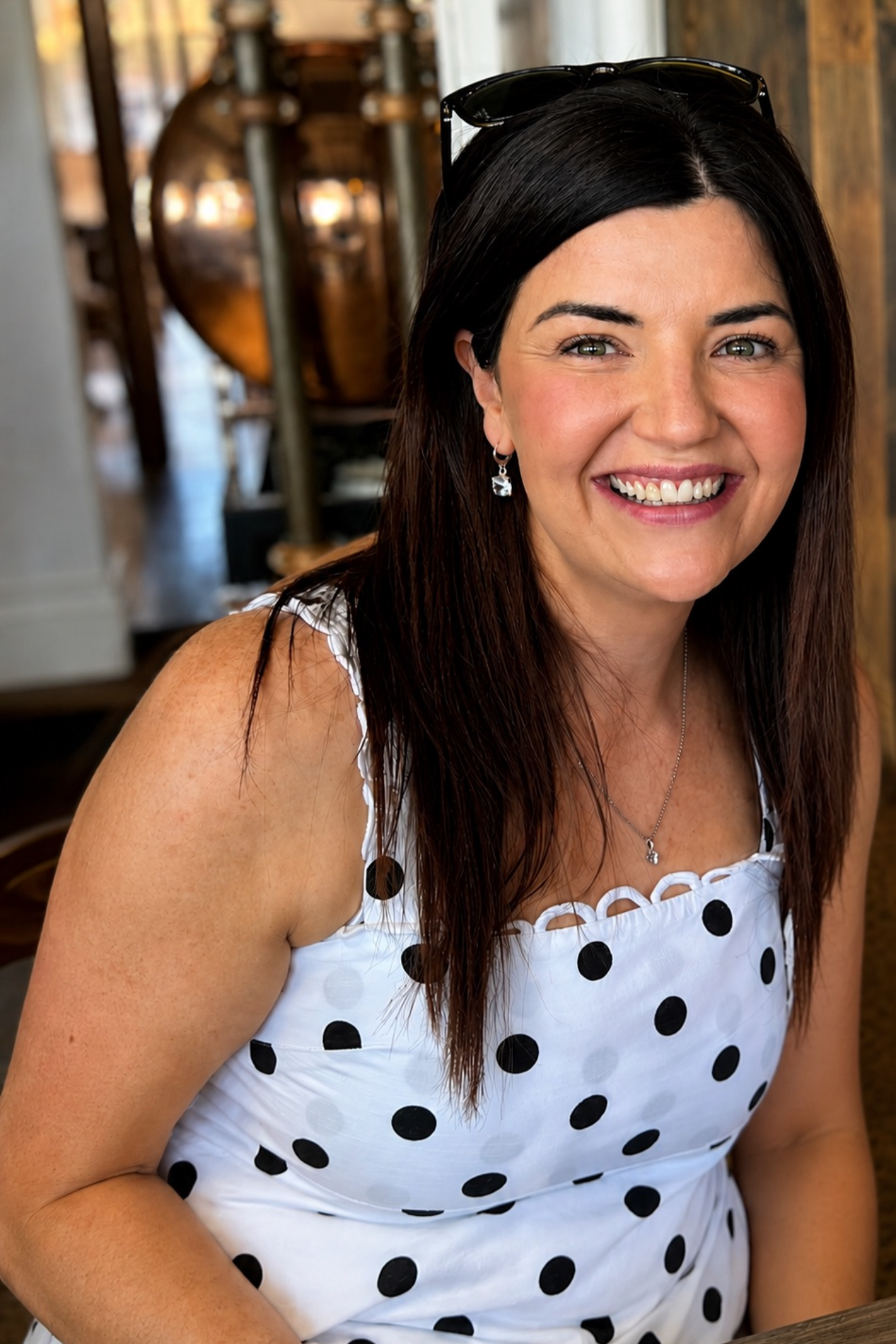 A woman with dark brown hair, wearing earrings and sunglasses on her head, smiling at the camera. She has a fair complexion with freckles and is dressed in a white dress with black polka dots. The background appears to be an indoor setting with wooden and metallic decor.
