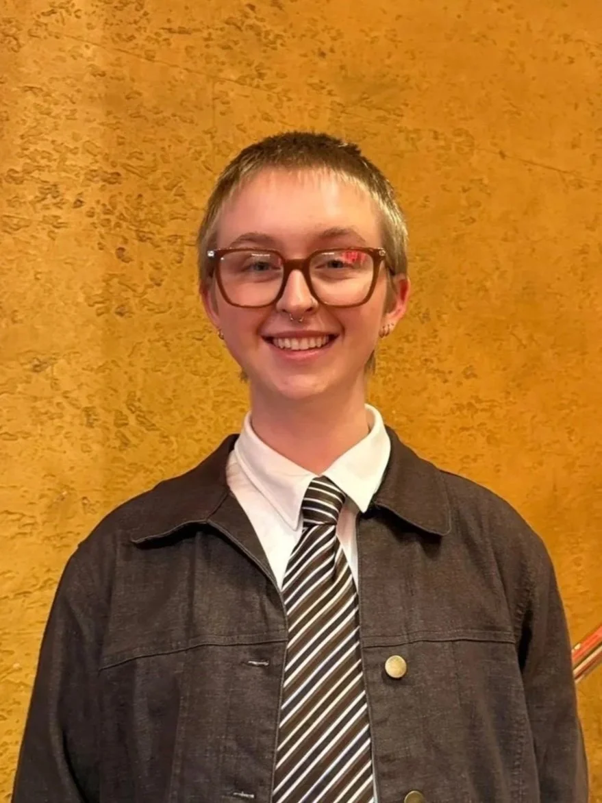 Person sitting at a restaurant table with a plate, wearing glasses and a black outfit, with a brick wall background.