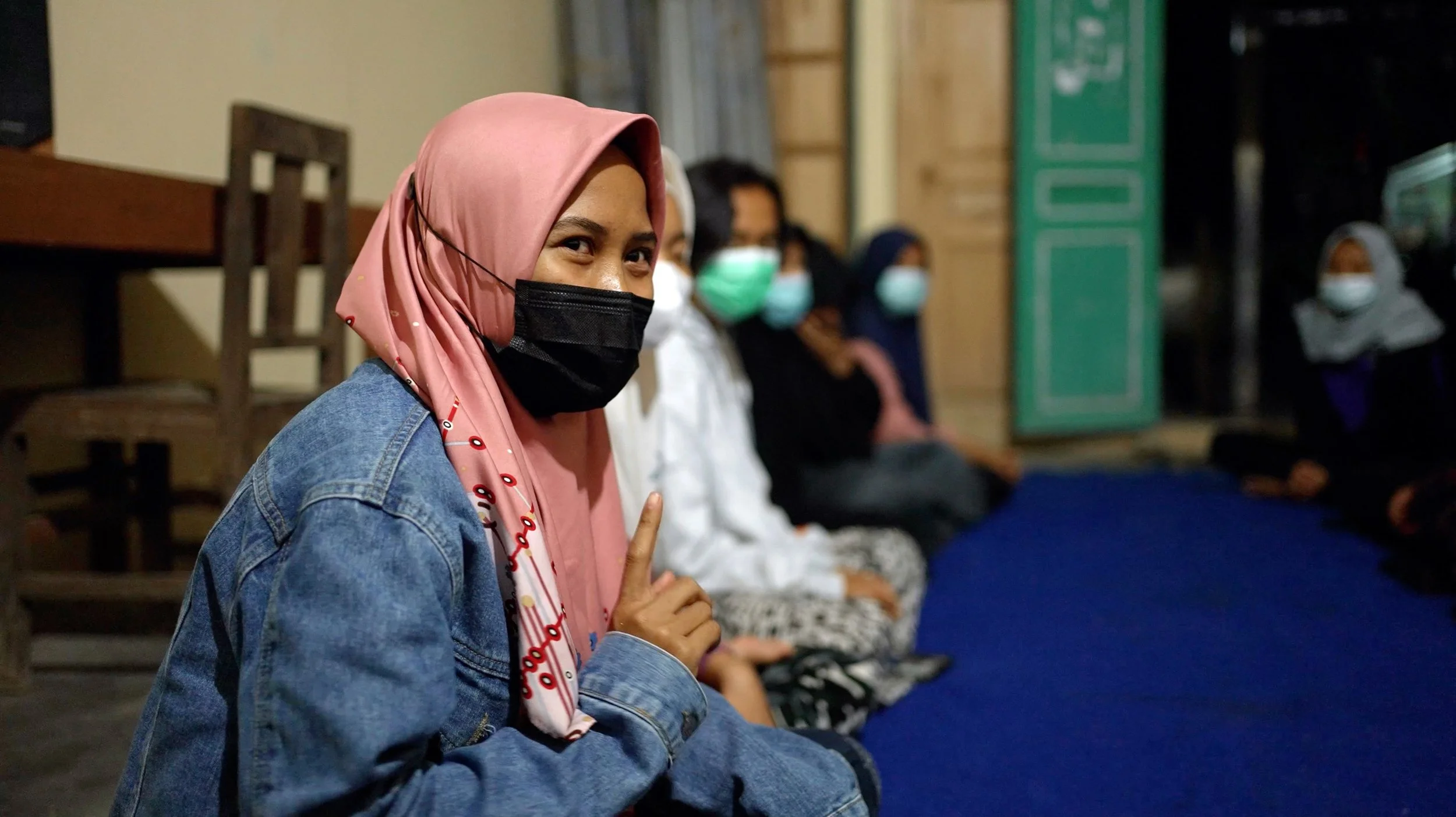 A woman wearing a pink hijab, black face mask, and denim jacket, sitting indoors on a blue carpet with her index finger raised, with several other women sitting nearby, all wearing face masks.