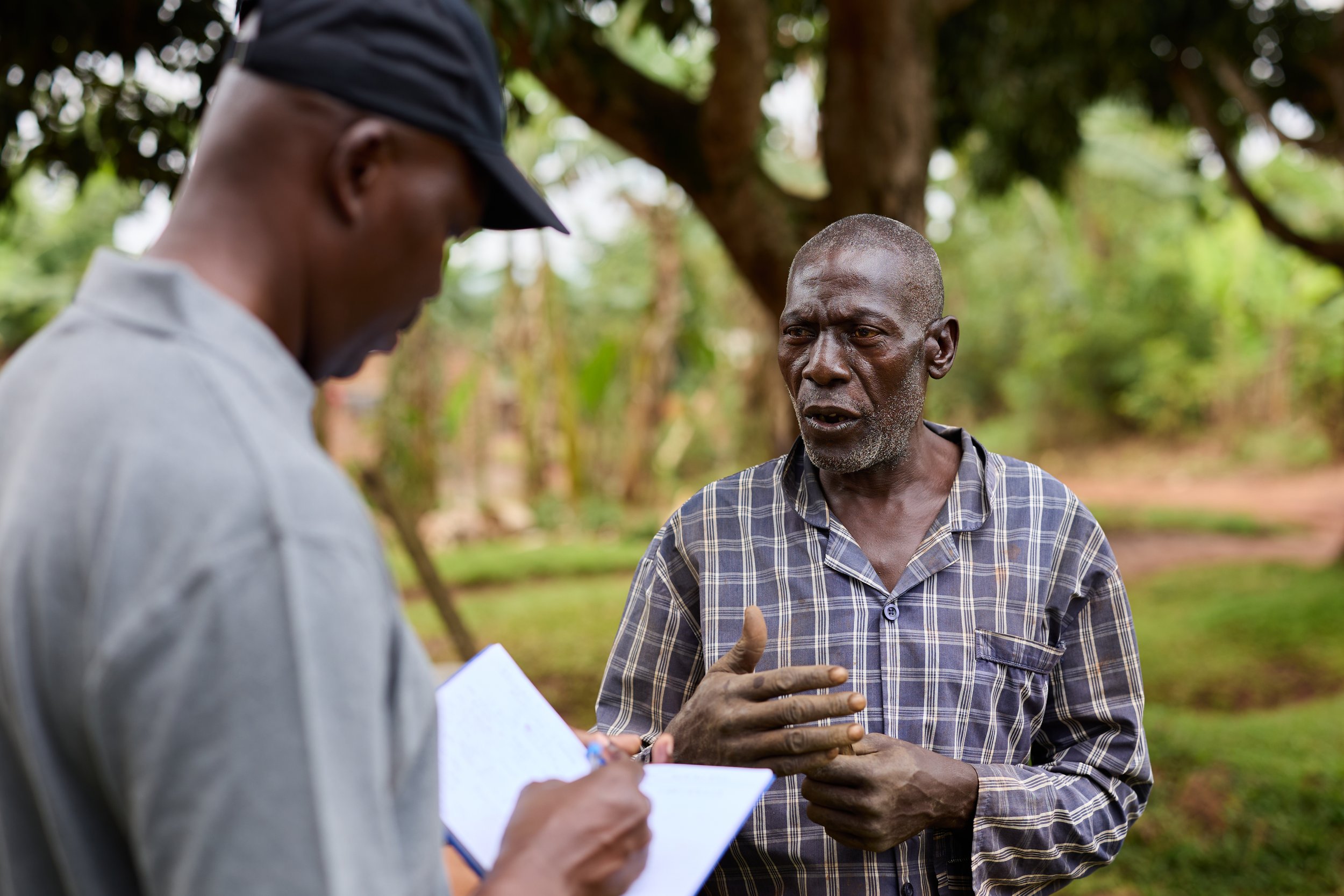 Outdoor scene with two men in conversation, one is writing in a notebook and the other is speaking with a serious expression, surrounded by trees and greenery.