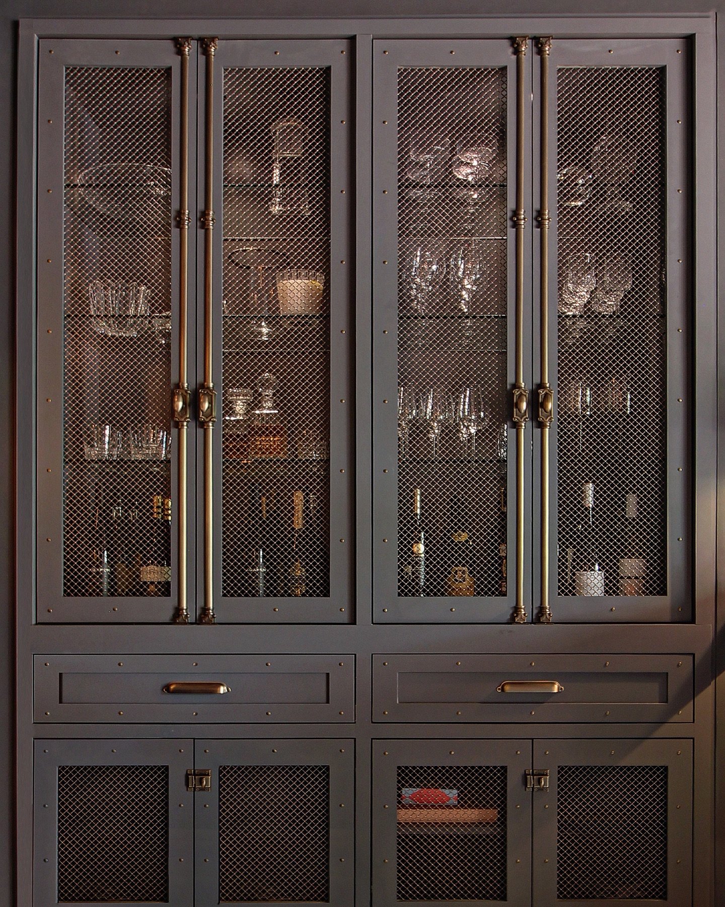Hardware is where design whispers luxury.

In this bespoke cabinet, every detail lends itself to the overall: from antique brass Cremone bolts to color-matched rivets and a delicate bronze metal mesh revealing crystal, glassware, and spirits. A contr